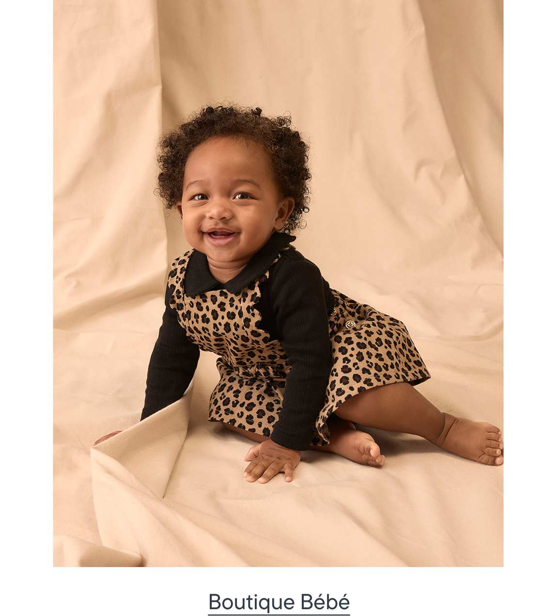 Un bébé souriant, vêtu d'une robe à imprimé léopard, est assis sur un fond en tissu beige.