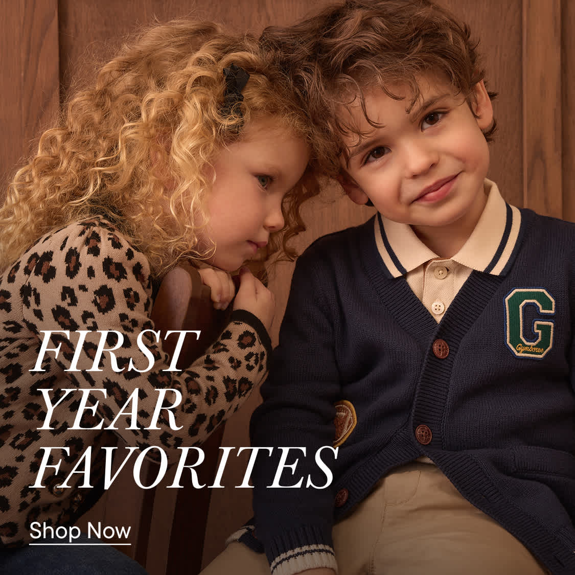 Two young children, a girl with curly blonde hair and a boy with brown hair, are leaning against each other in front of a wooden background, with the text "FIRST YEAR FAVORITES" overlaid on the left side.