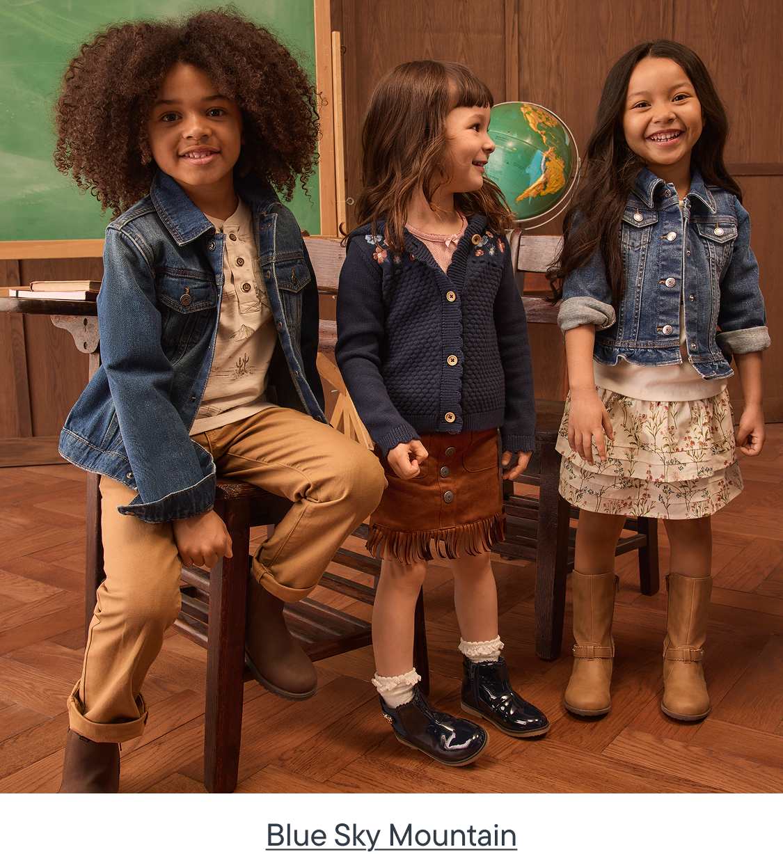 Three children in casual outfits pose in a classroom setting with a globe and chalkboard in the background.