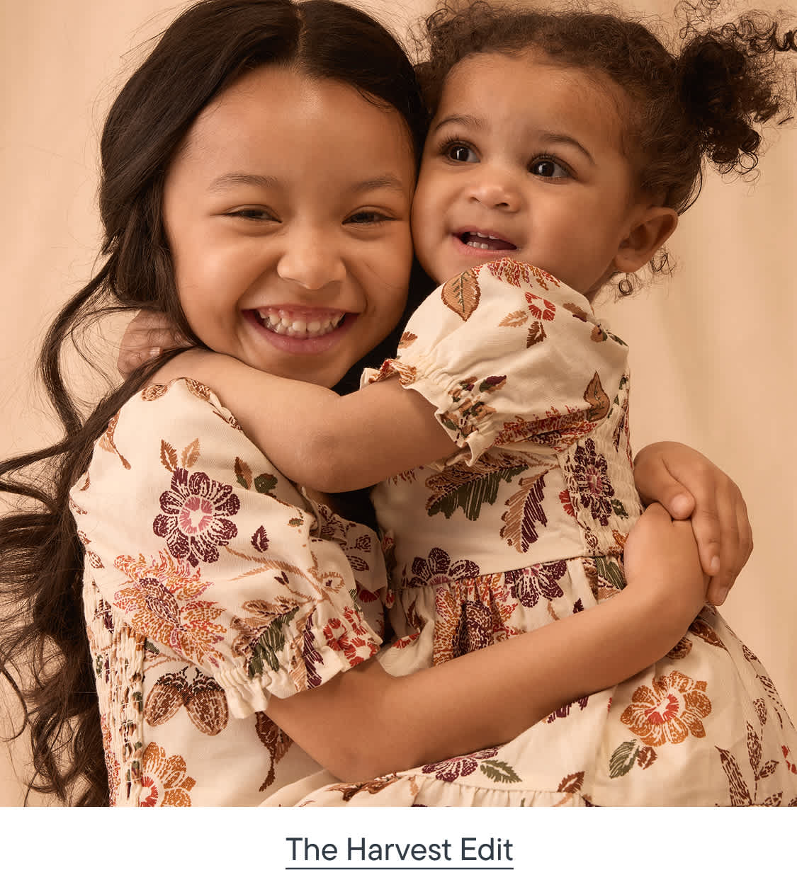 Two young girls in floral dresses embrace each other against a soft, neutral background.