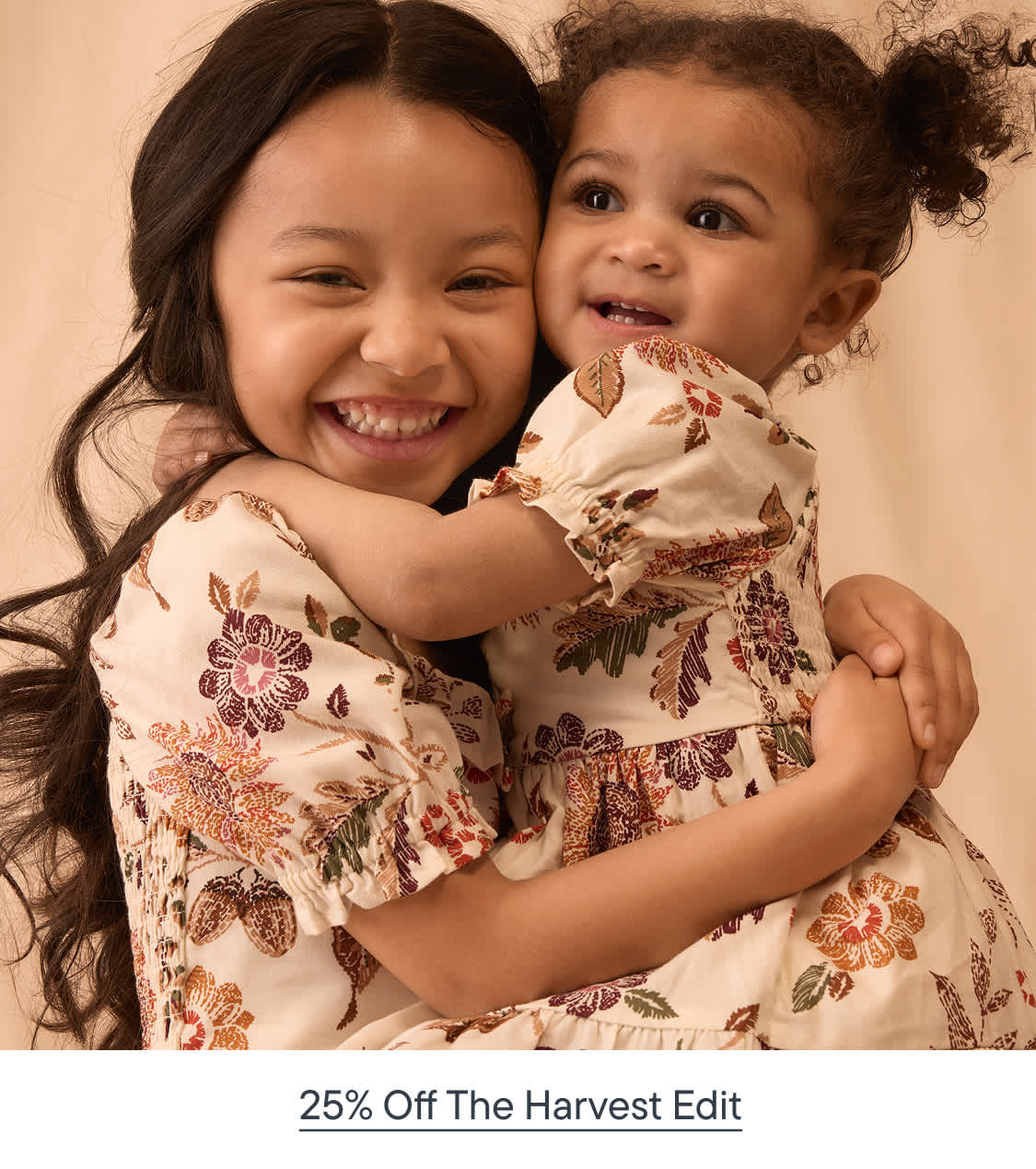 Two young girls in floral dresses hug each other against a plain, neutral-toned background.