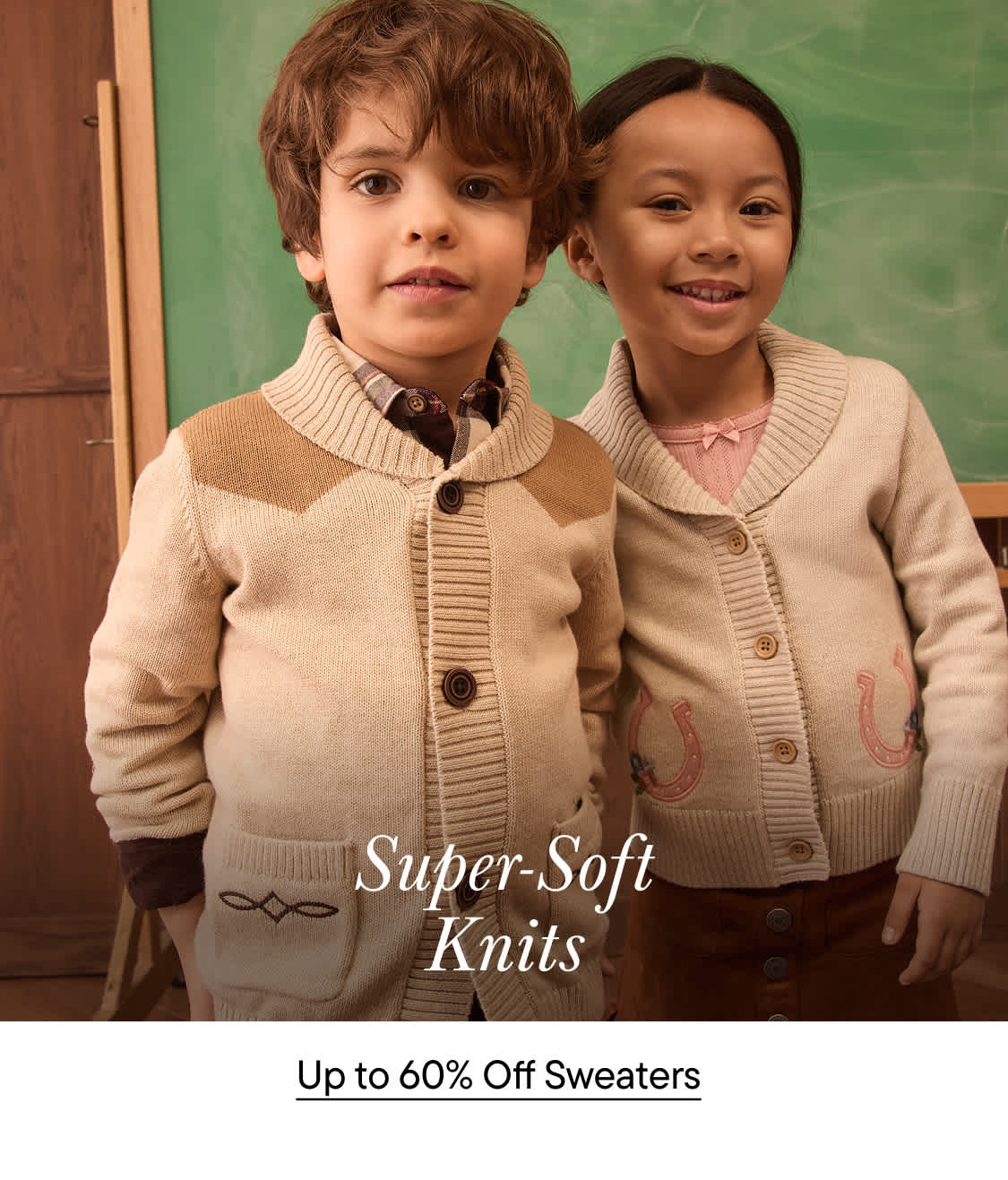 Two young children wearing beige knit cardigans smile in front of a green chalkboard.