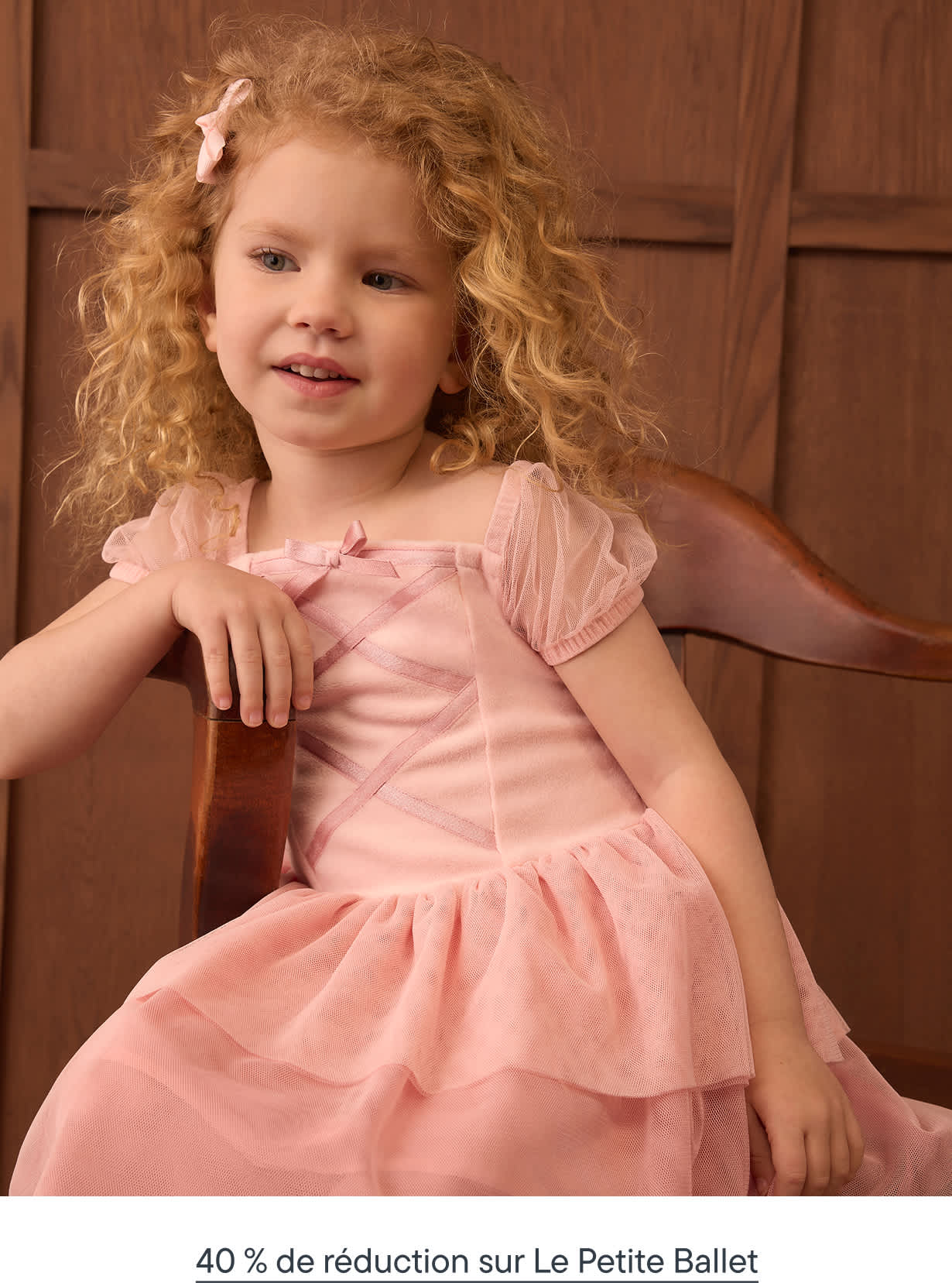 Une jeune fille aux cheveux blonds bouclés, vêtue d'une robe rose, est assise sur une chaise en bois contre un mur lambrissé marron.