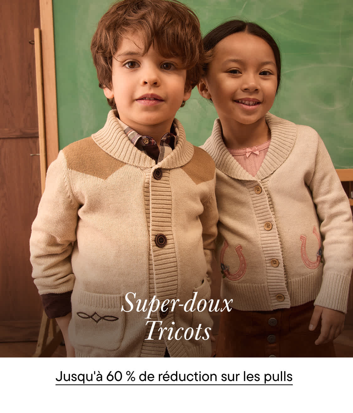 Deux jeunes enfants vêtus de gilets en tricot beige sourient devant un tableau noir vert.