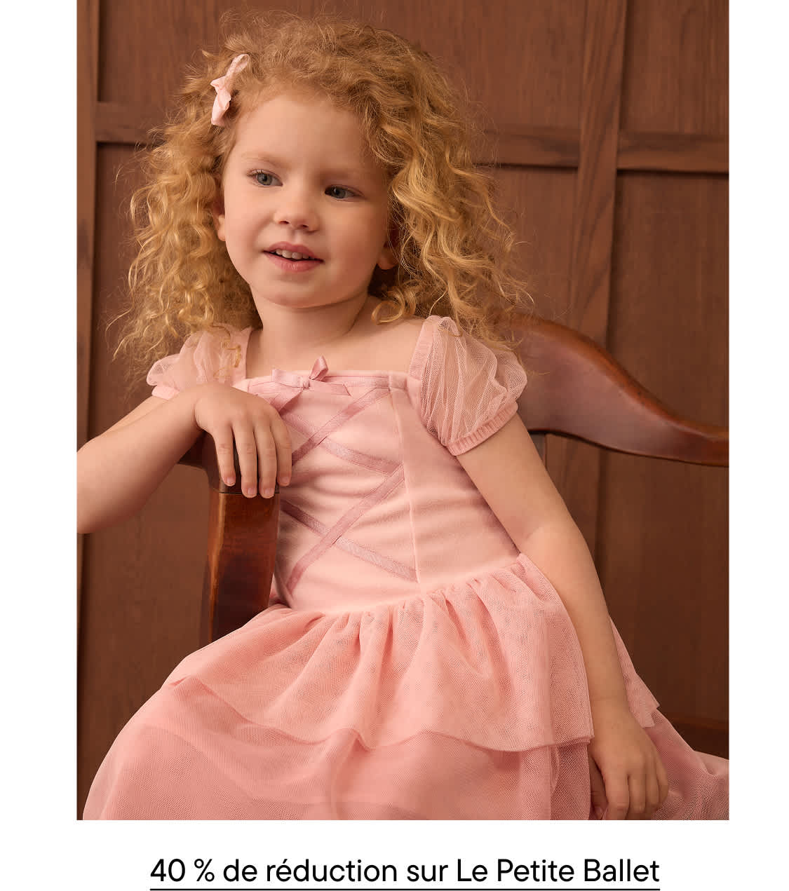 Une jeune fille aux cheveux blonds bouclés, vêtue d'une robe rose, est assise sur une chaise en bois contre un mur lambrissé marron.