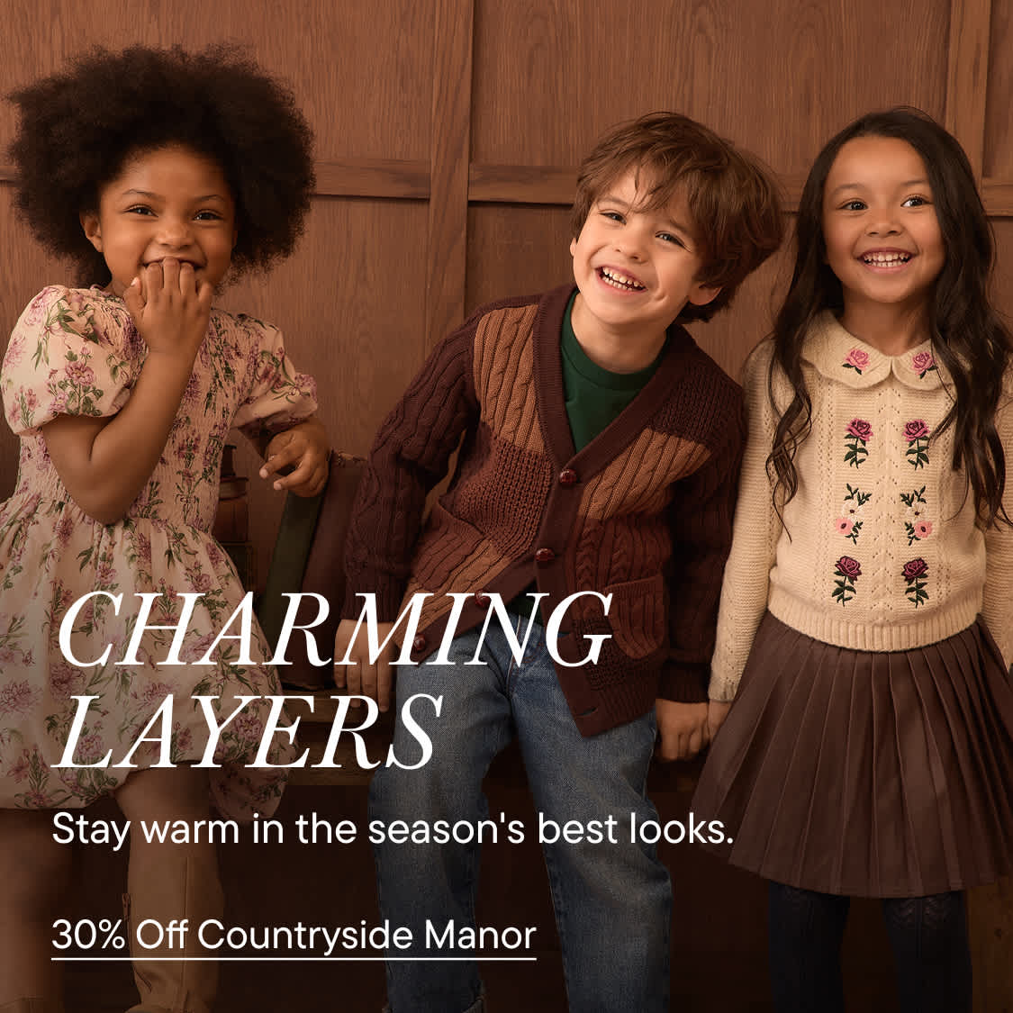 Three smiling children in autumn clothing stand in the foreground against a backdrop of brown wooden paneling.