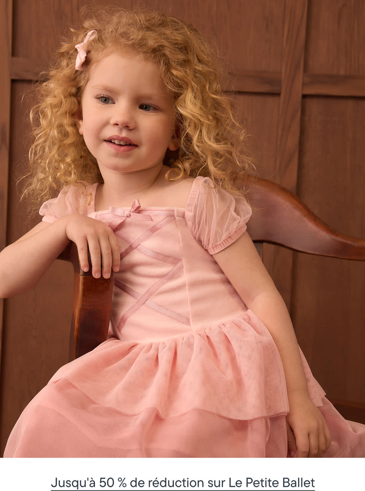 A young girl with curly blonde hair wearing a pink dress sits in a wooden chair against a paneled brown wall.