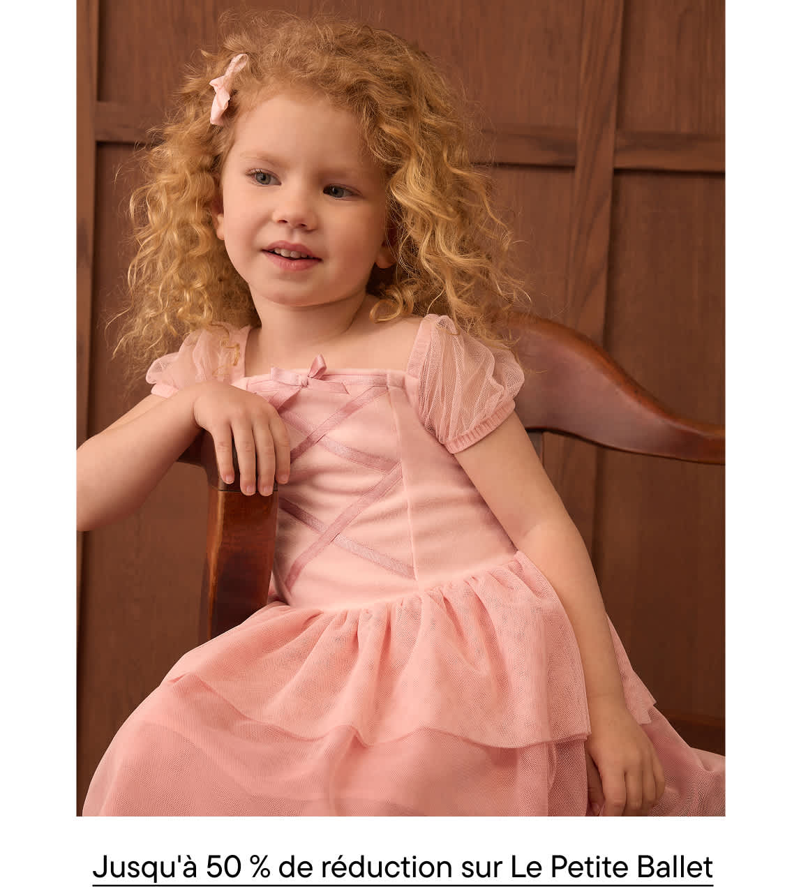 A young girl with curly blonde hair wearing a pink dress sits in a wooden chair against a brown paneled wall.