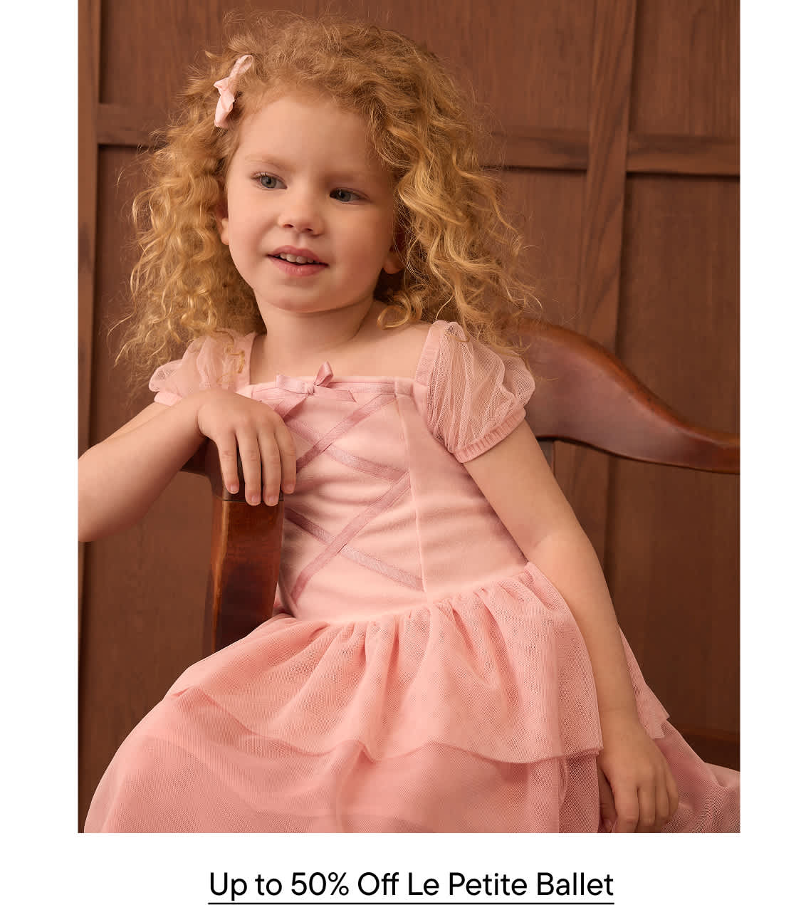 A young girl with curly blonde hair wearing a pink dress sits in a wooden chair against a brown paneled wall.