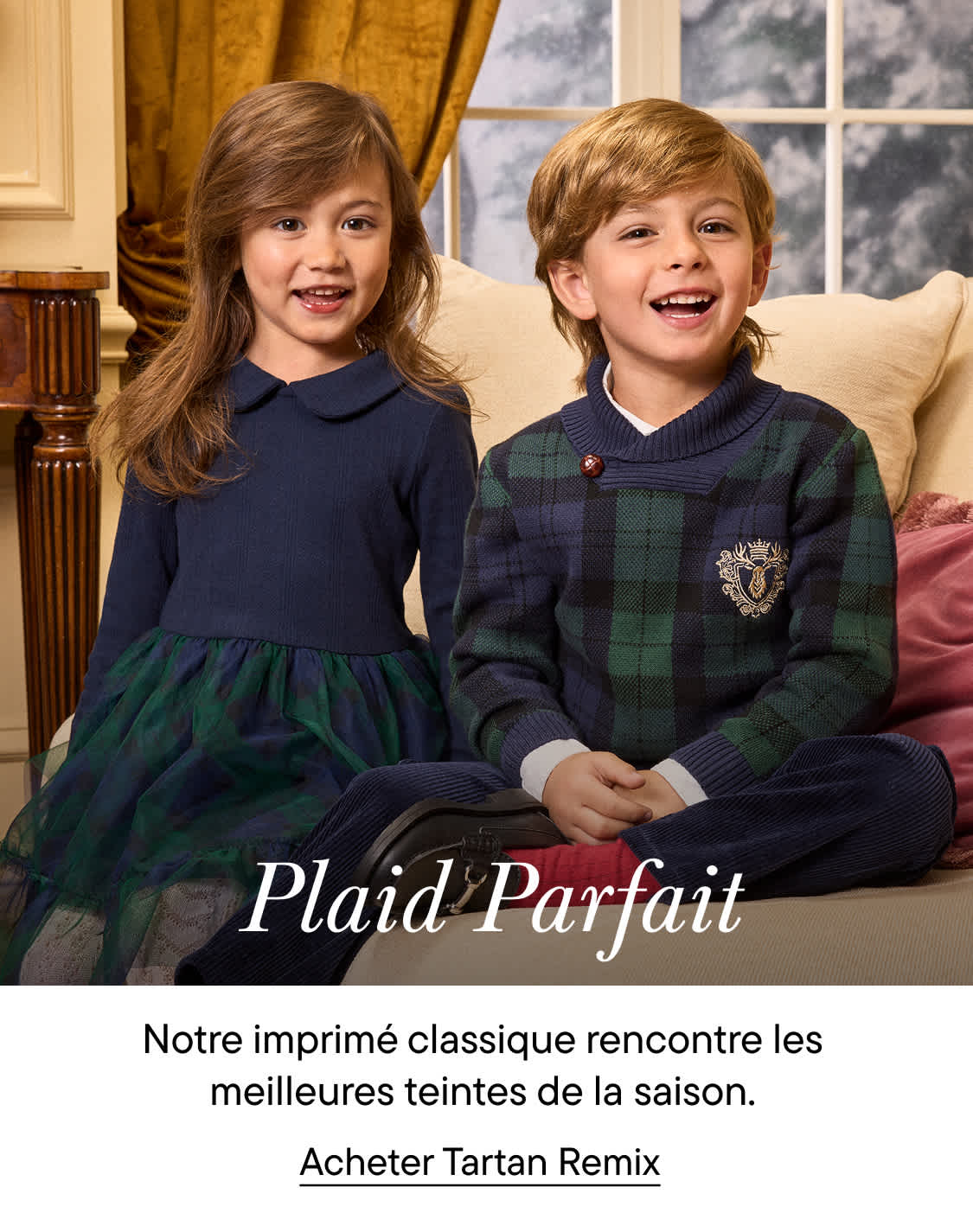 Deux enfants souriants, vêtus de vêtements bleu marine et à carreaux, sont assis sur un canapé beige dans une pièce meublée de façon classique.