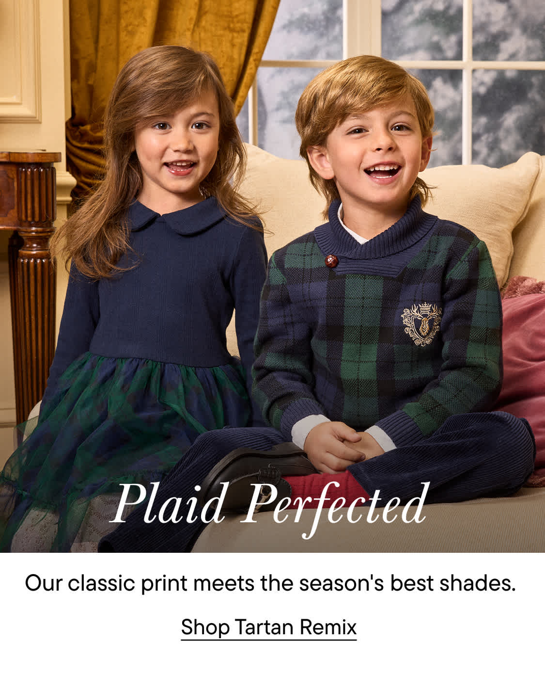 Two smiling children in navy and plaid clothing sit on a beige sofa in a classically furnished room.