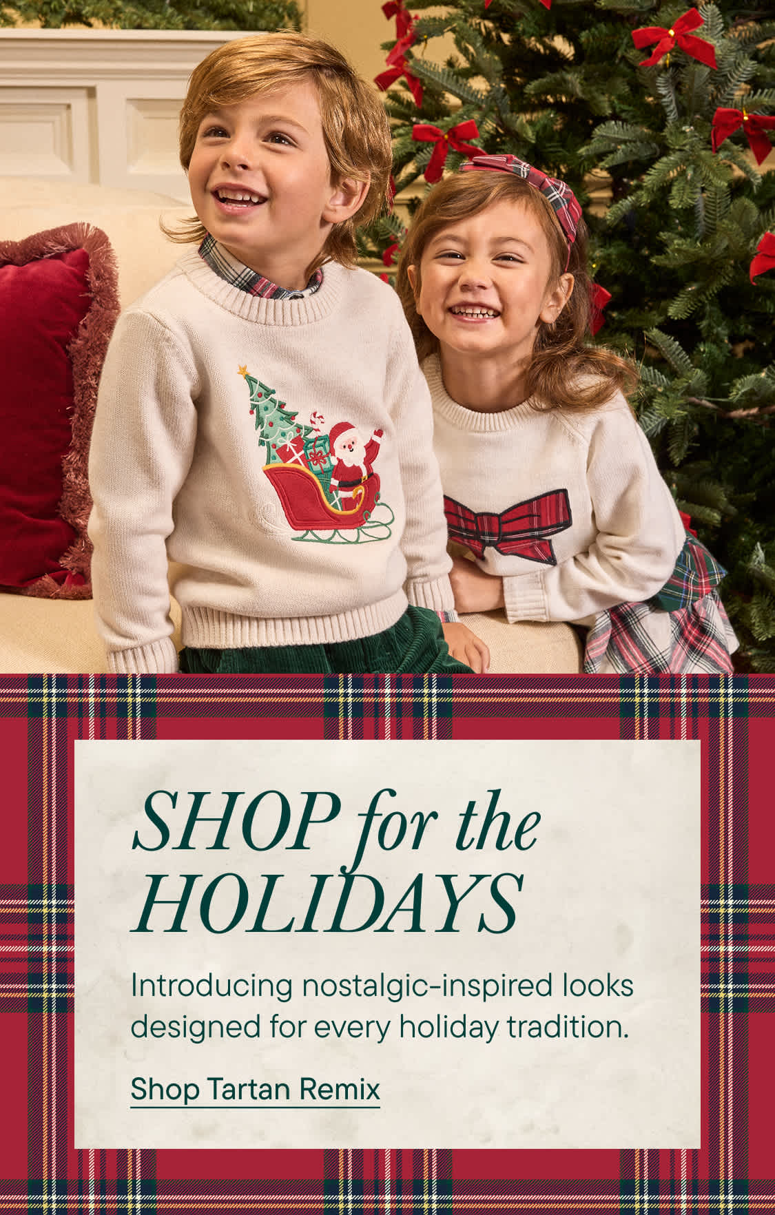 Two smiling children in holiday sweaters sit in the foreground, with a decorated Christmas tree and a cozy room in the background.