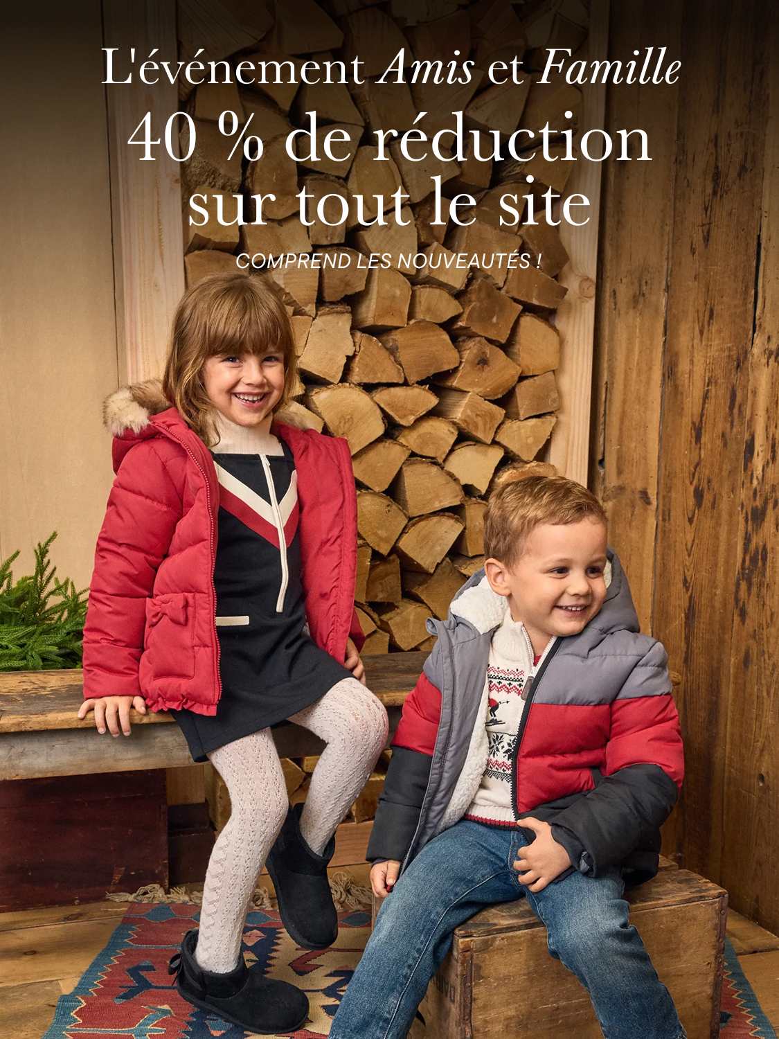 Deux jeunes enfants en doudounes sourient au premier plan, devant un tas de bois de chauffage empilé et un mur en bois.