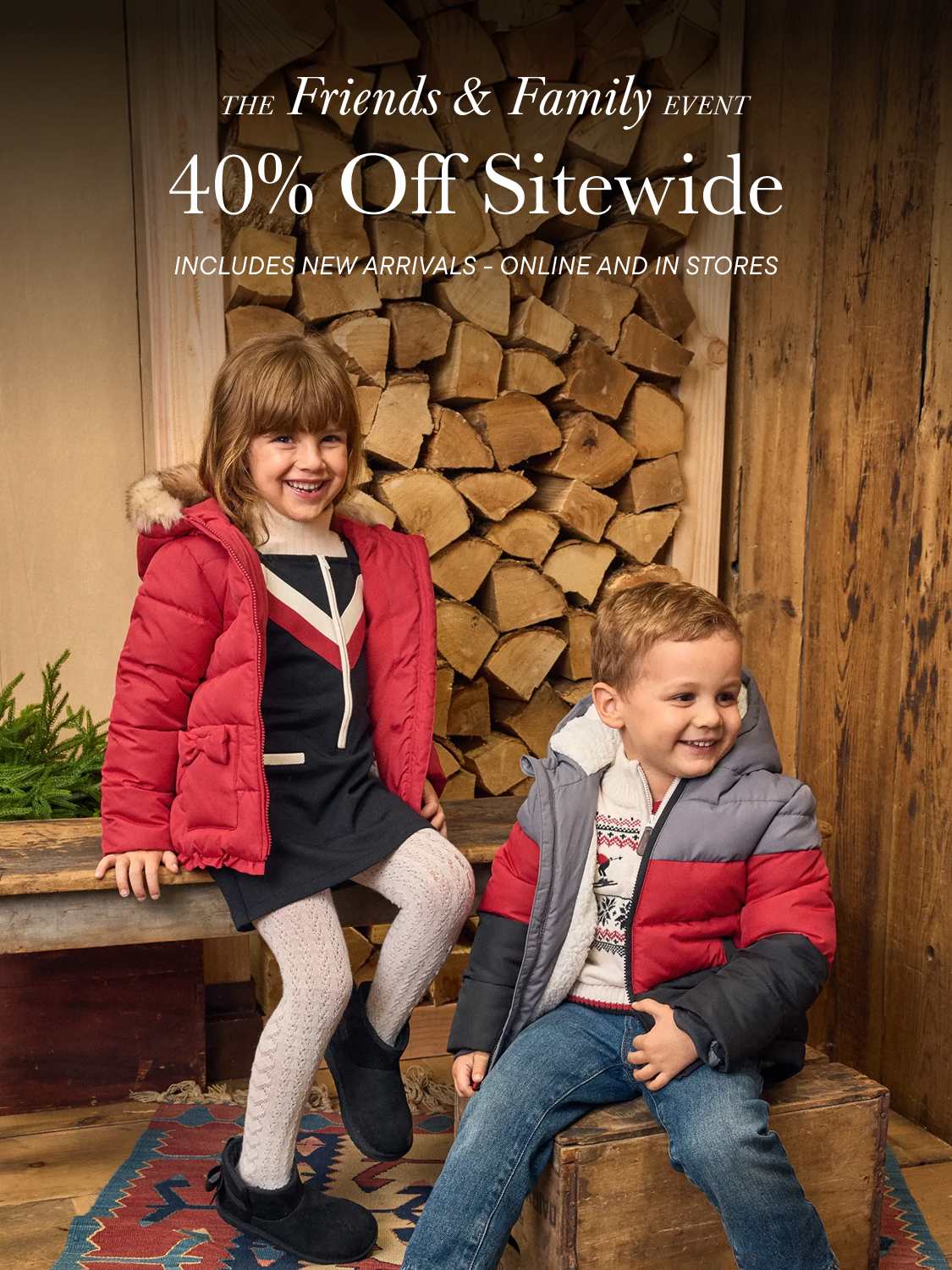 Dos niños pequeños con chaquetas de invierno sonríen en primer plano, con un fondo de leña apilada y una pared de madera.