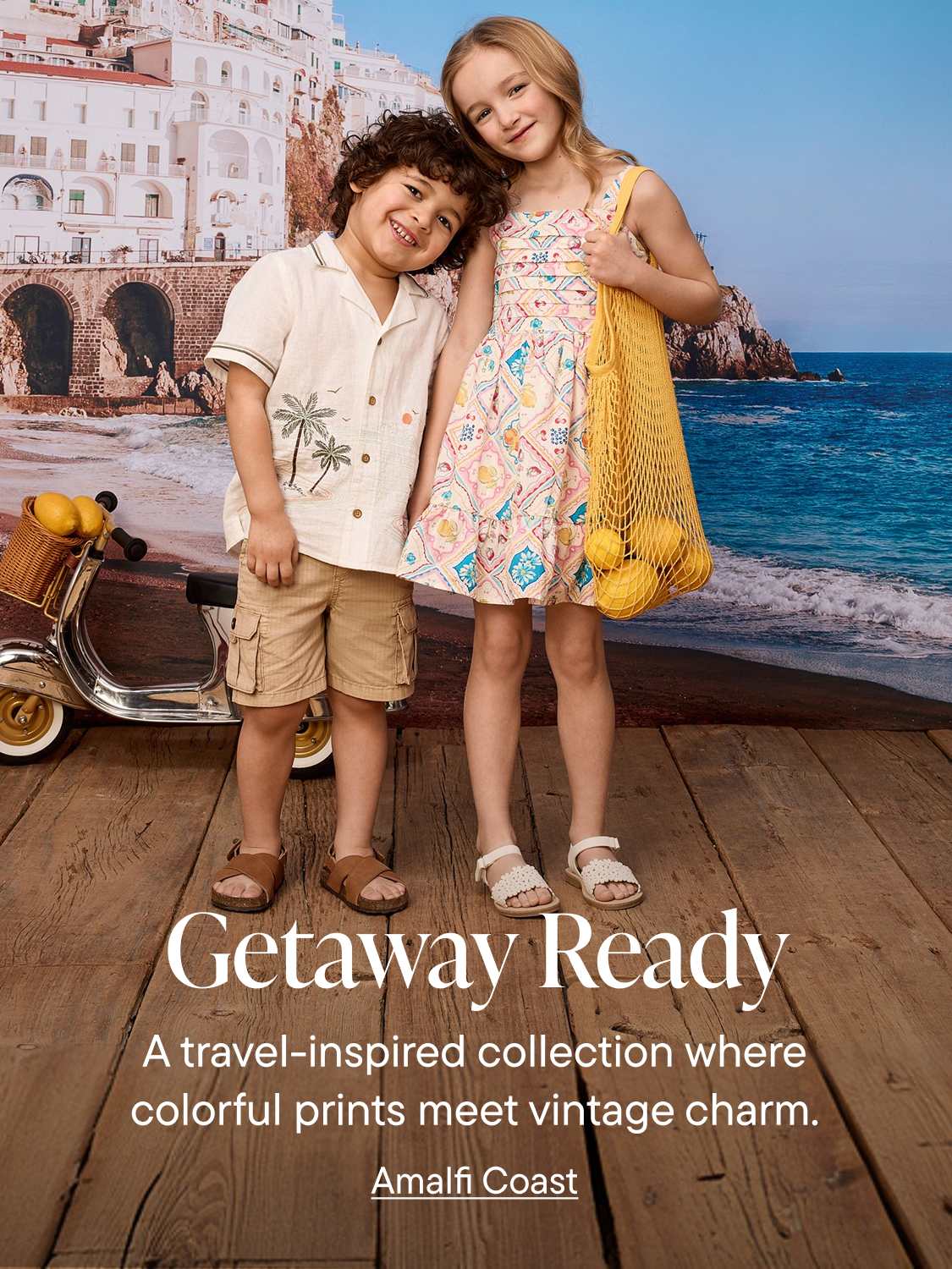 Two children stand on a wooden deck in front of a backdrop of a coastal town and the ocean, with a scooter to the left.