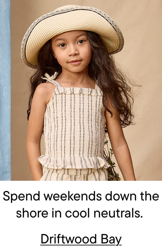 A young girl in a straw hat and striped outfit stands against a neutral background with a hint of blue fabric on the left.