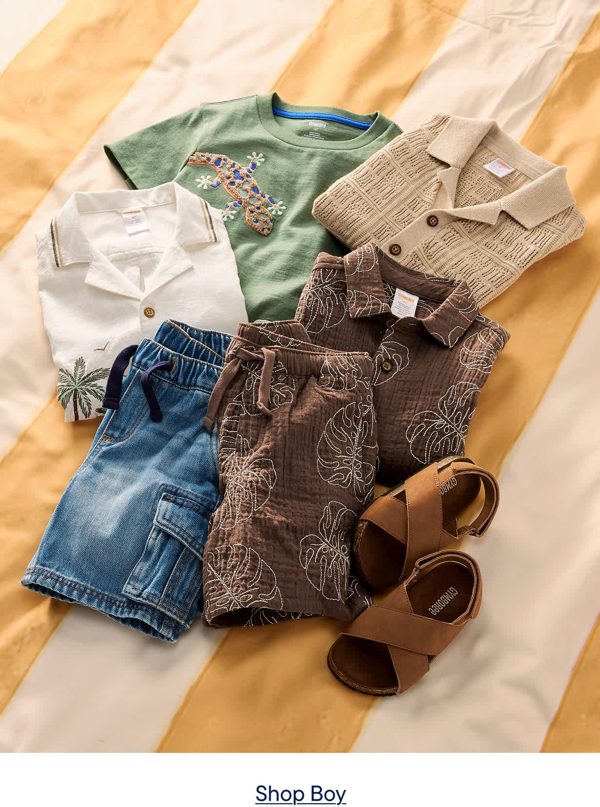 A collection of boys' summer clothes, including shorts, shirts, and sandals, are laid out on a striped yellow and white surface.