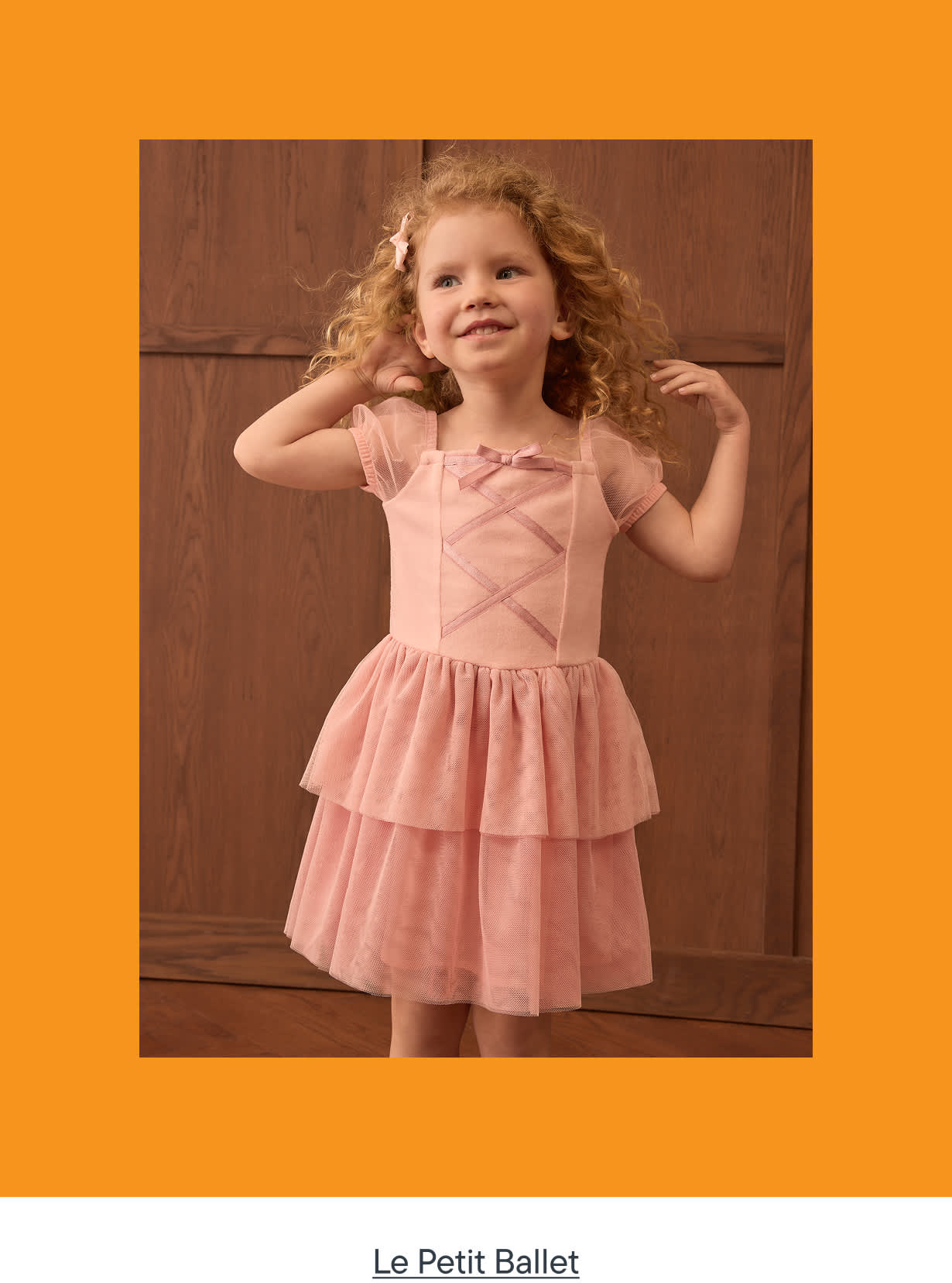 A young girl with curly blonde hair smiles as she poses in a pink ballet dress with a tiered skirt, standing in front of a wooden door.