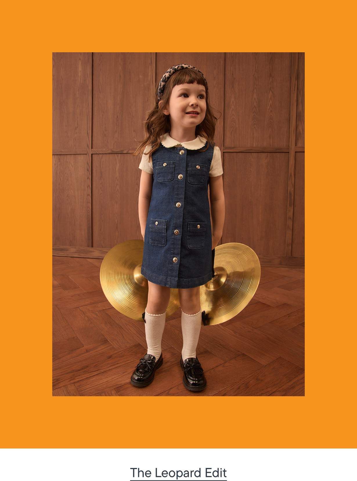 A young girl in a denim dress and beret stands in front of two large cymbals against a wood-paneled wall.