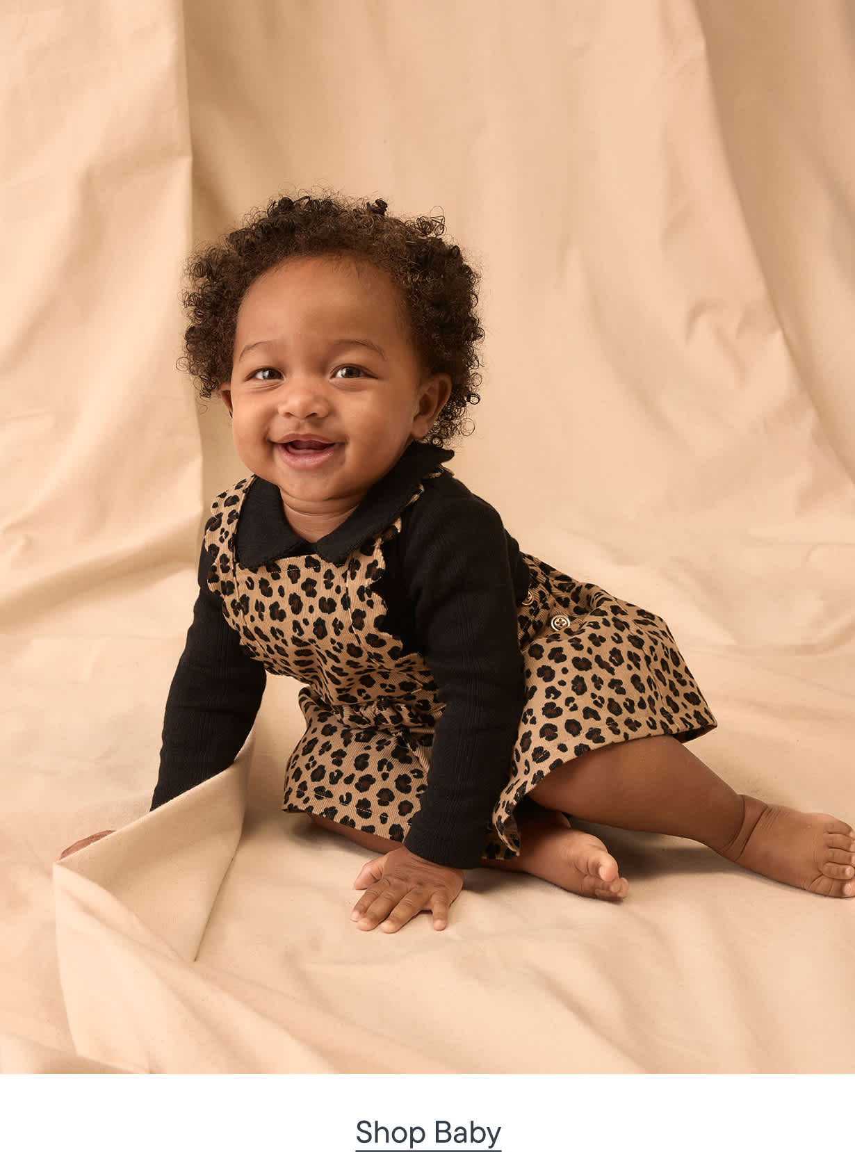 A smiling baby in a leopard print dress sits on a cream-colored fabric backdrop.