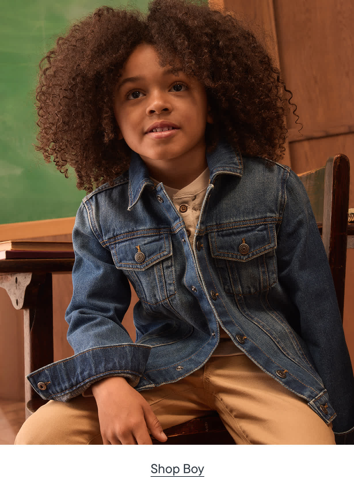 A young boy with curly hair, wearing a denim jacket and tan pants, sits in front of a chalkboard.