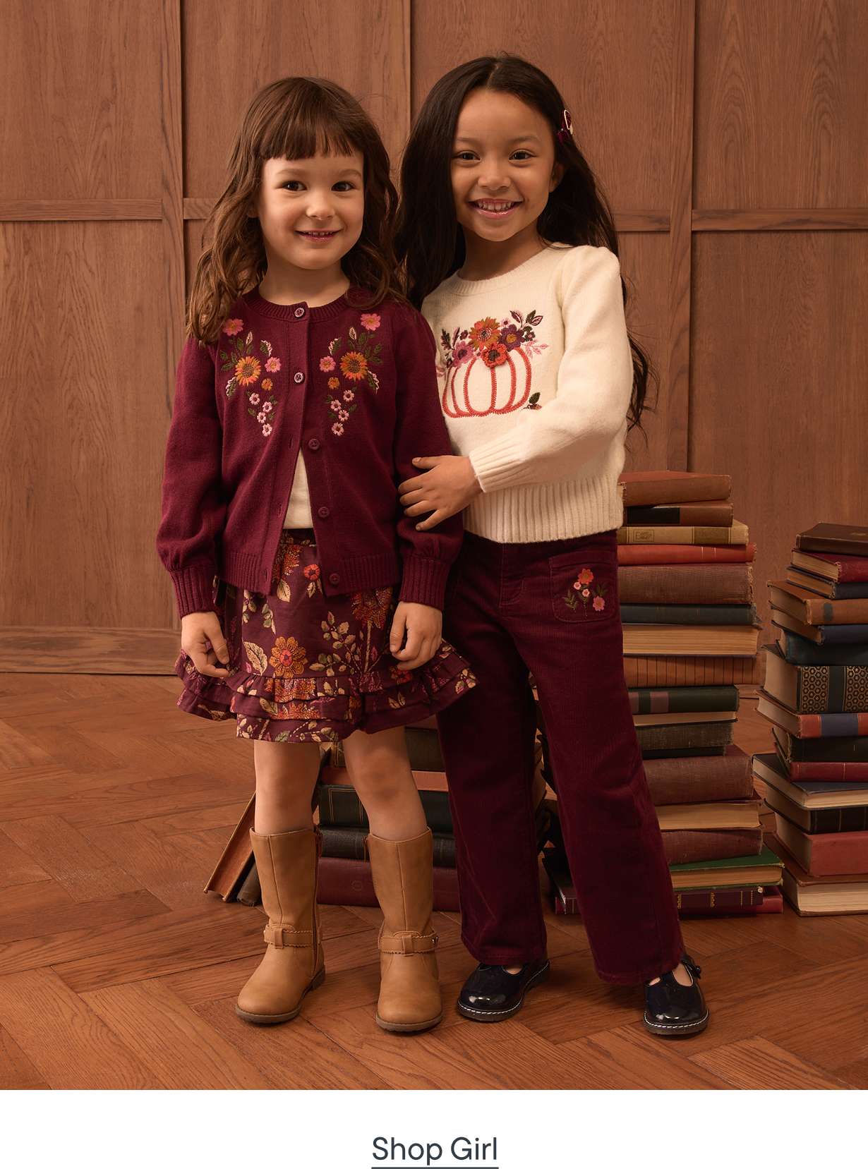 Two young girls in autumnal outfits stand in front of a stack of books and a wooden wall.