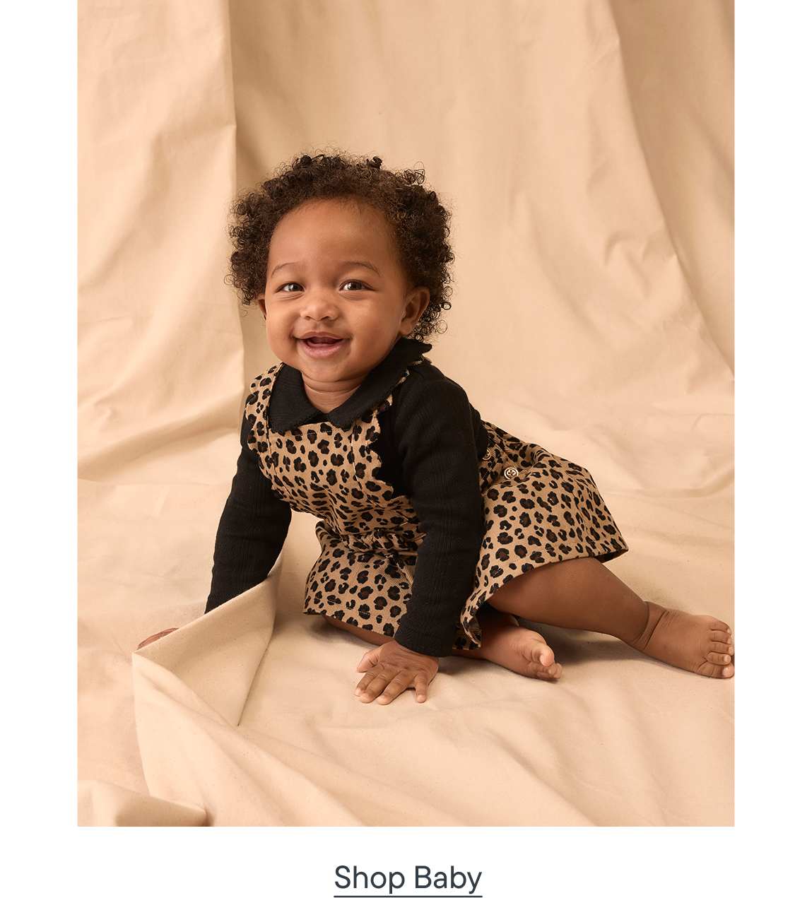 A smiling baby in a leopard print dress sits on a beige fabric background.
