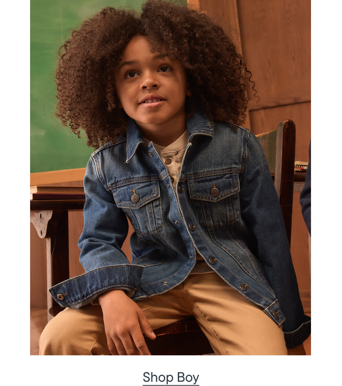 A young boy with curly hair, wearing a denim jacket and tan pants, sits in front of a classroom setting with a chalkboard and wooden desk.
