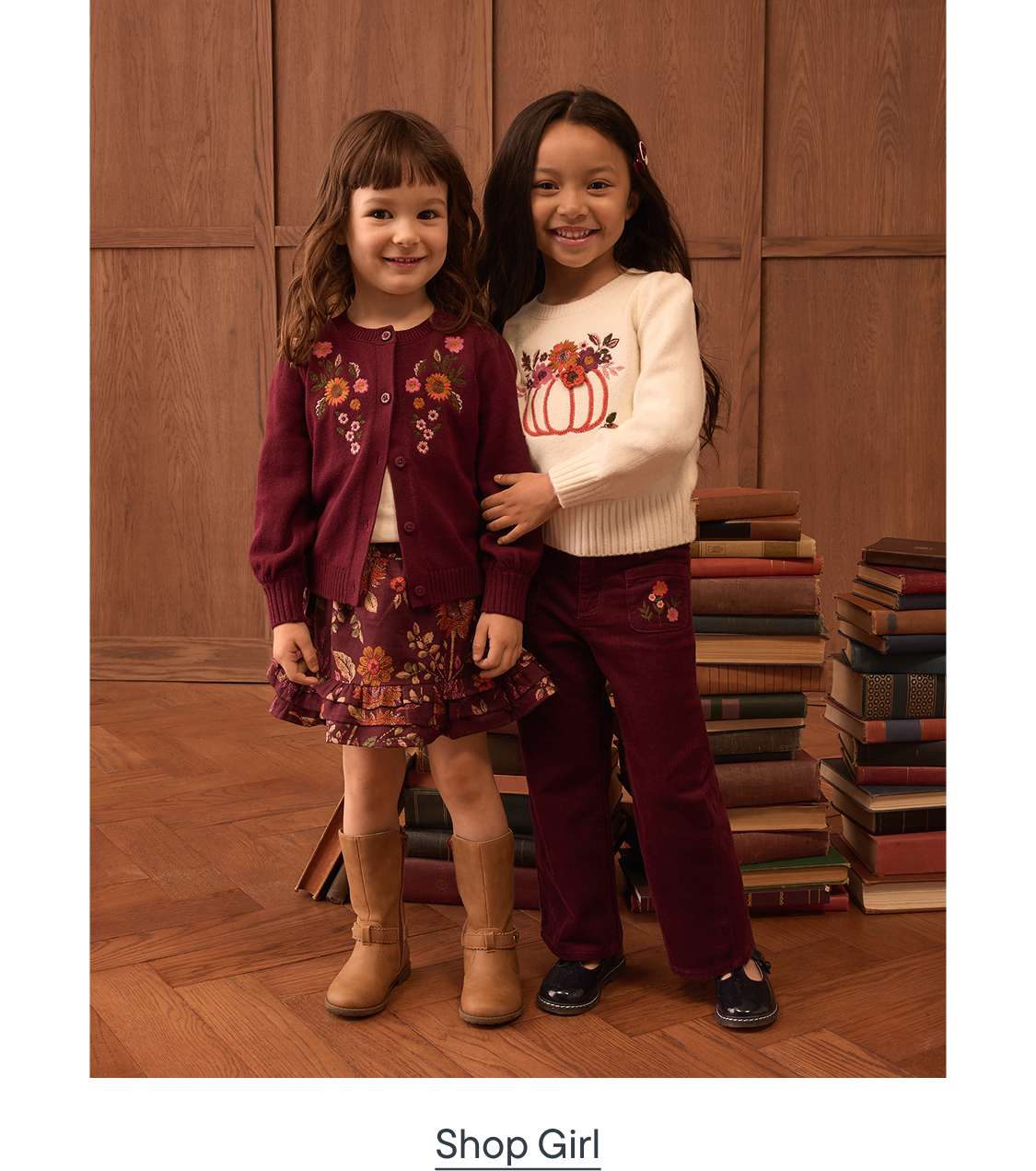 Two young girls in autumnal outfits stand in front of a stack of books and a wooden wall.