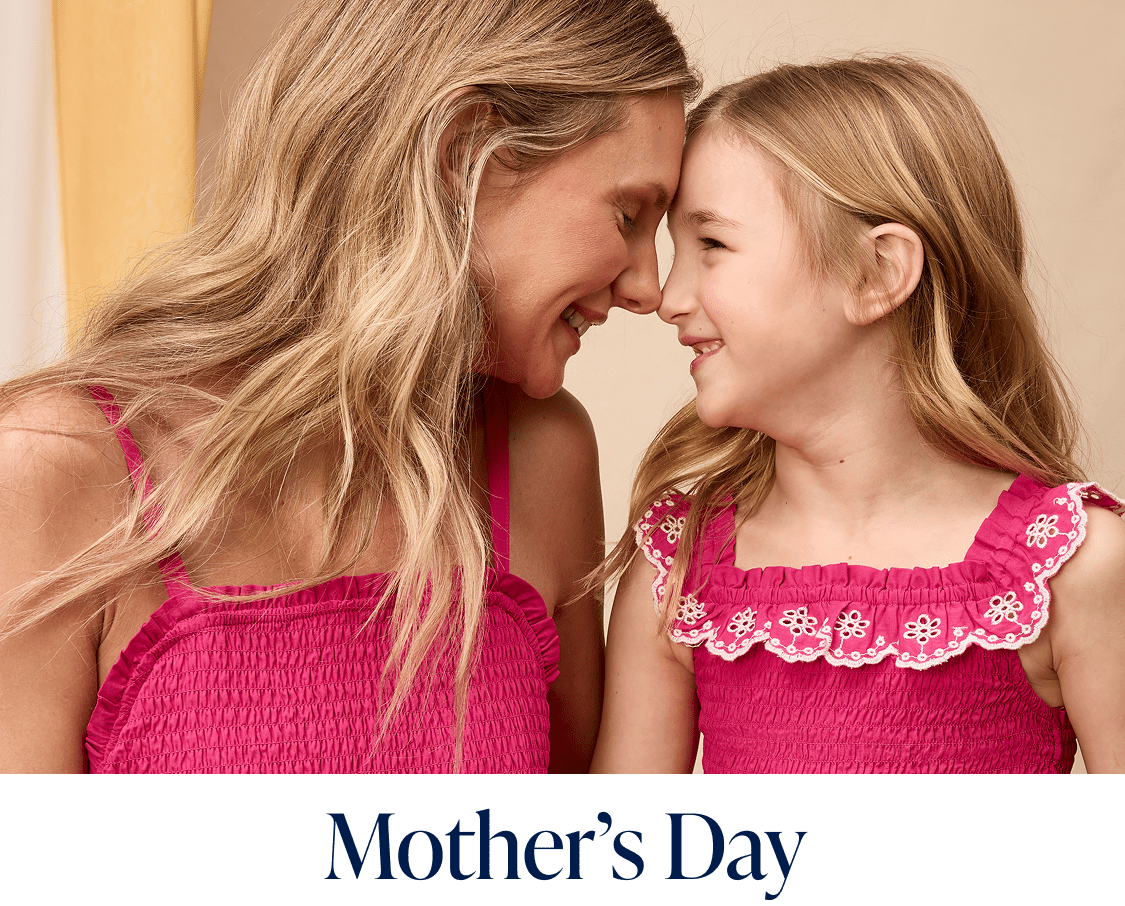 Two young girls with long blonde hair wearing matching pink dresses are smiling and interacting with each other in what appears to be a warm and affectionate manner.