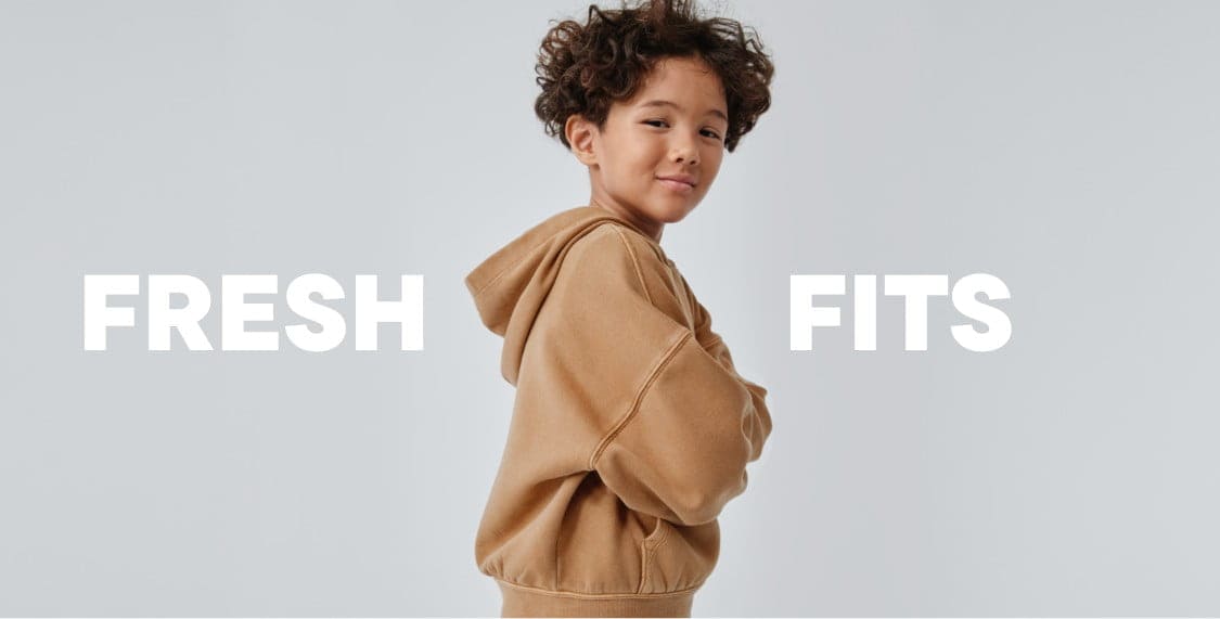A young person with curly hair wearing a beige sweater stands against a plain white background with the text "FRESH FITS" prominently displayed.