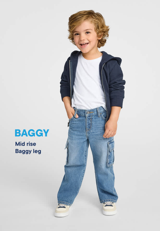 Un niño pequeño con una sudadera azul con capucha y vaqueros está de pie sobre un fondo blanco liso, con la palabra "BAGGY" y "Mid rise Baggy leg" escritas en texto azul a su izquierda.