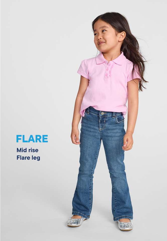 A young girl in a pink polo shirt and flared jeans stands against a plain white background.