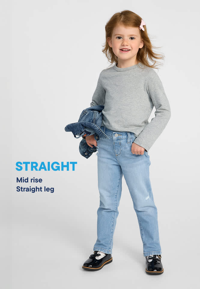 A young girl with red hair and a pink bow stands in the foreground wearing a grey long-sleeved shirt and light blue jeans, holding a denim jacket, against a plain white background.