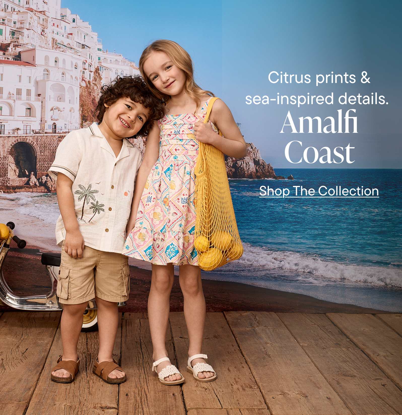 The image features two young people, a man and a woman, standing together on a wooden boardwalk with the Amalfi Coast and its colorful buildings visible in the background.