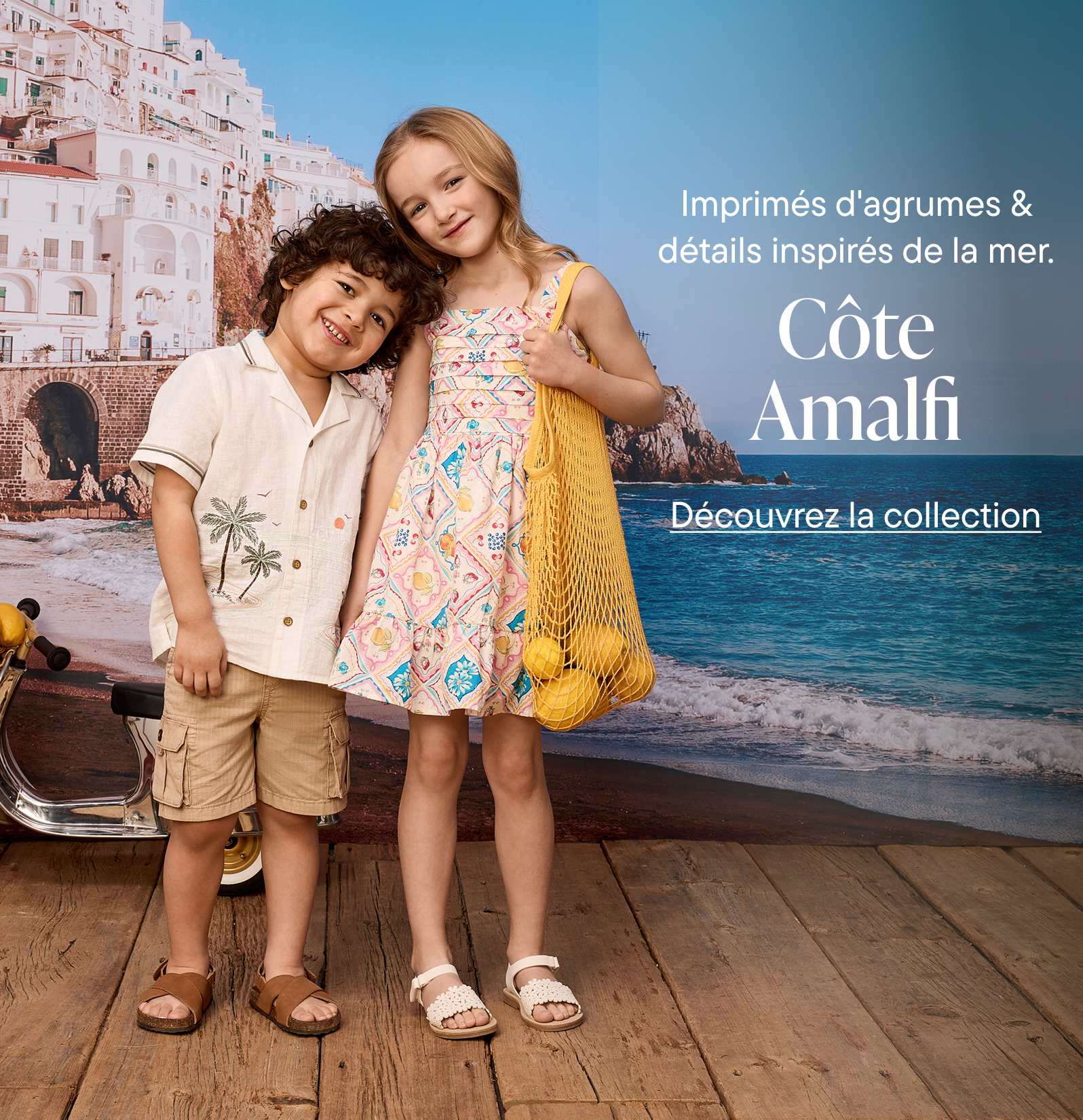 The image features two young people, a man and a woman, standing together on a wooden boardwalk with the Amalfi Coast and its colorful buildings visible in the background.