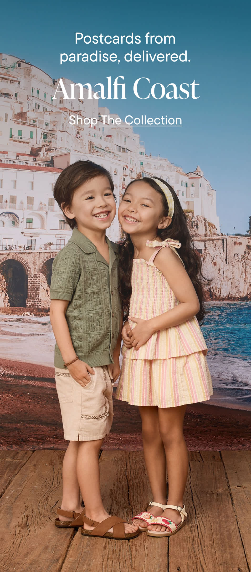 Two young children, a boy and a girl, are standing together in front of a scenic coastal town with colorful buildings and a body of water in the background. The children appear to be smiling and embracing each other, creating a warm and friendly atmosphere.