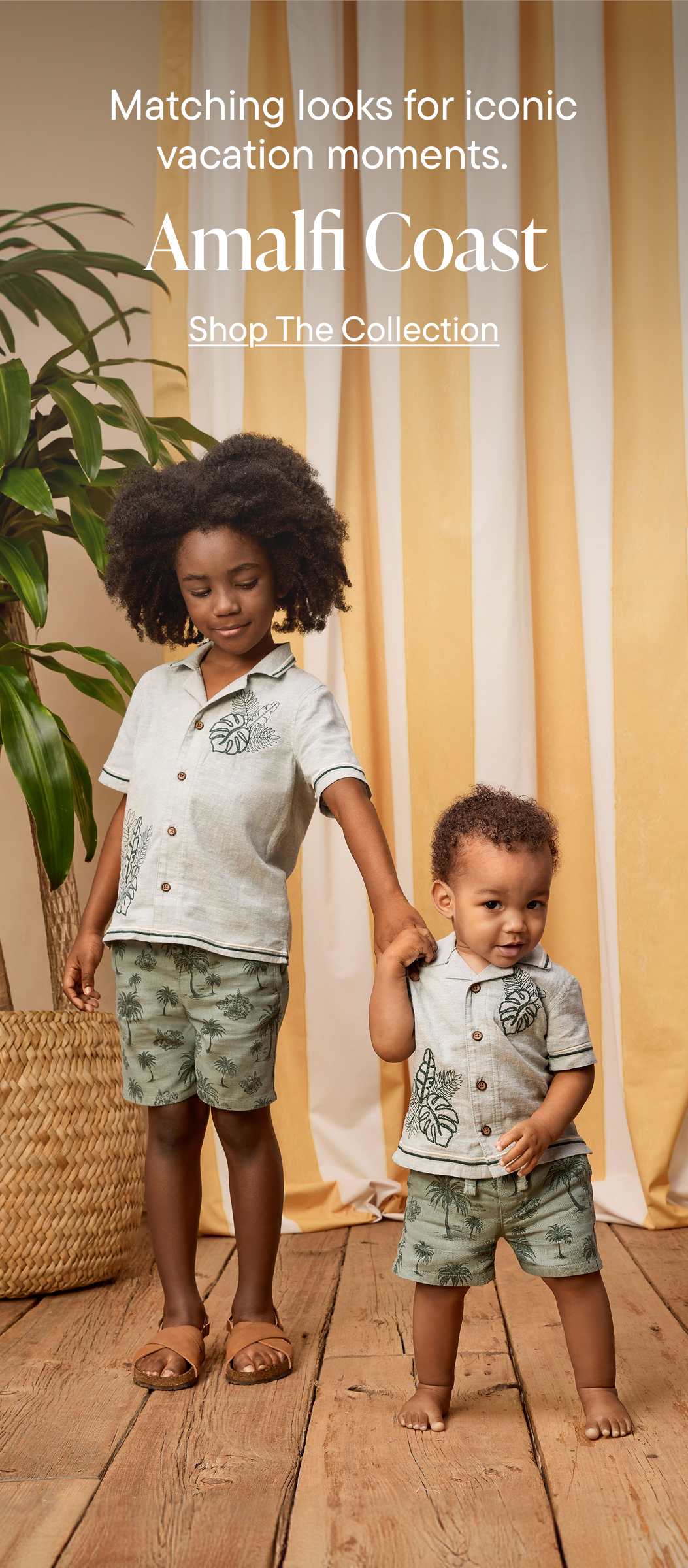 Two young children, a girl and a boy, are standing in front of a display for the Amalfi Coast clothing collection. The background features a tropical-themed backdrop with plants, suggesting a vacation or resort setting.