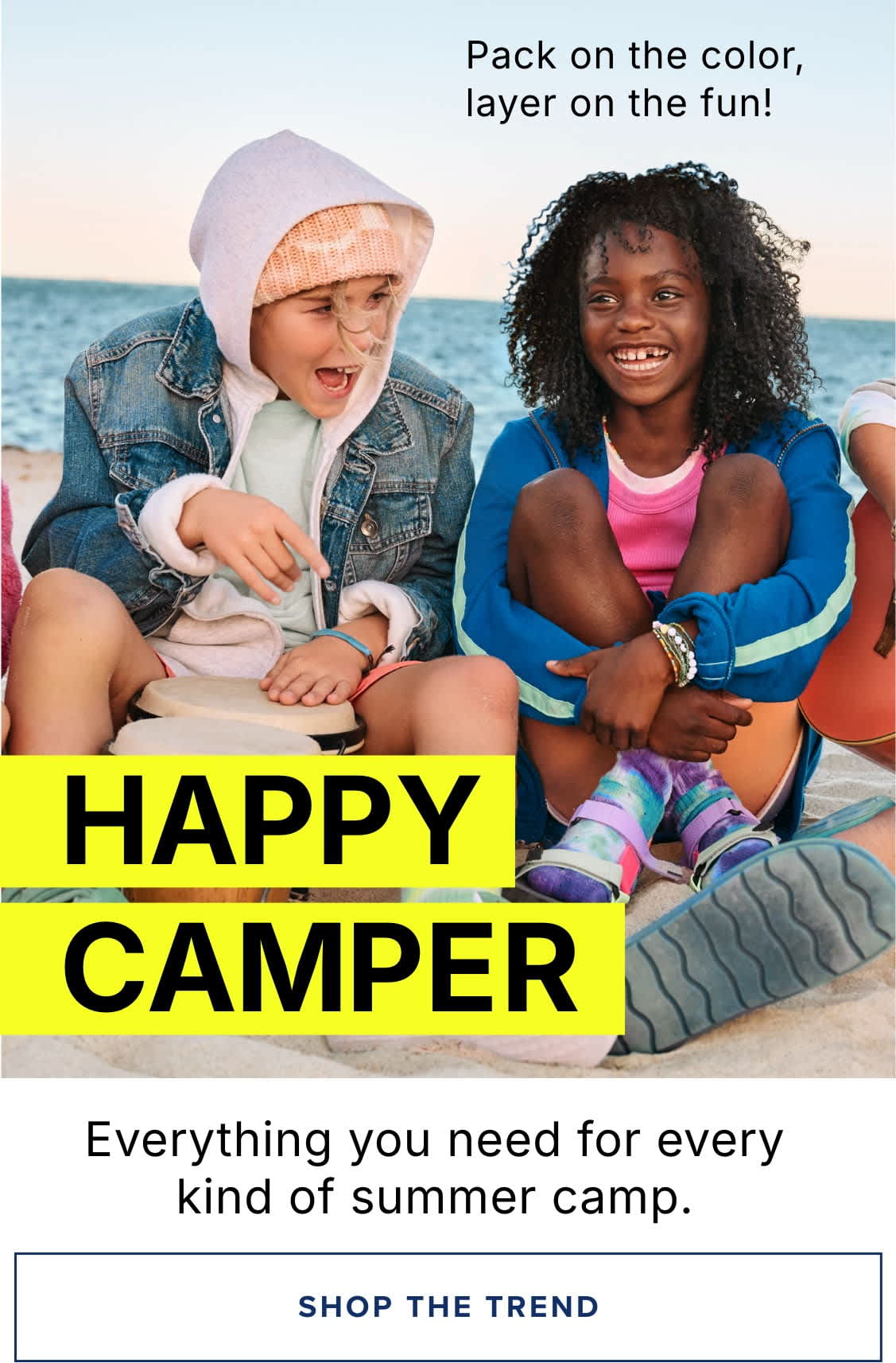 A group of young people, dressed in colorful and casual attire, are sitting on a beach, enjoying the outdoors and each other's company. The background features a scenic ocean view, suggesting a summer camp or outdoor adventure setting.