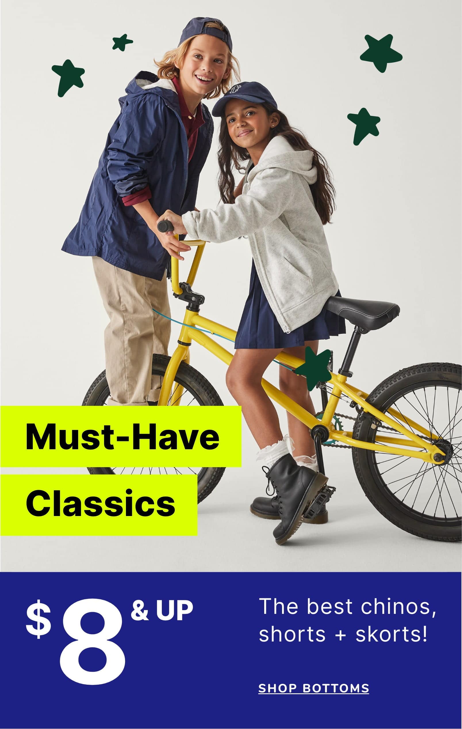 Two young girls, one with blonde hair and one with dark hair, are riding a yellow bicycle together against a plain background with green star shapes.