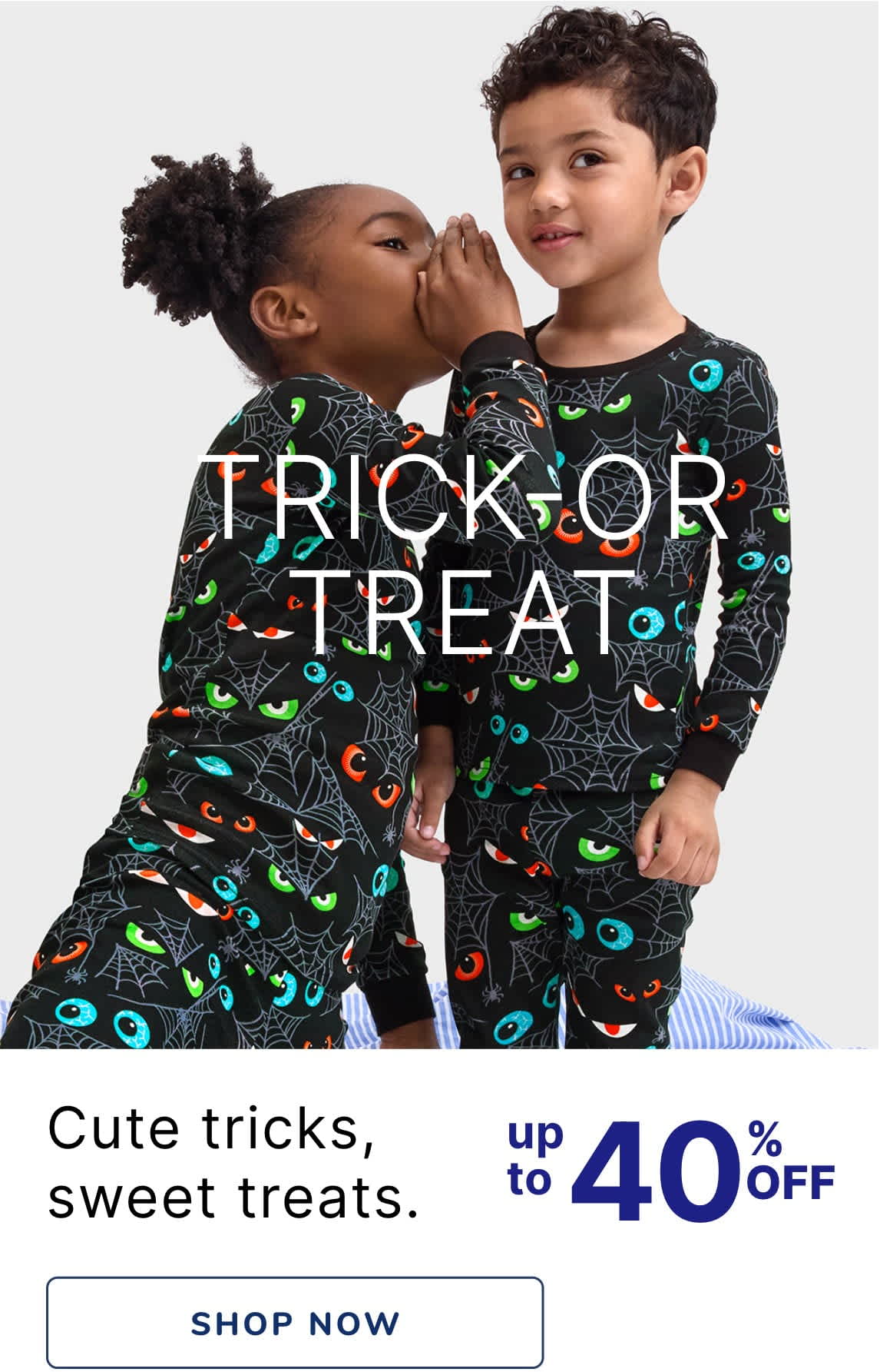 Two young children, a boy and a girl, wearing matching Halloween-themed pajamas and embracing each other affectionately. The background is a plain white backdrop, allowing the focus to remain on the children's playful interaction.
