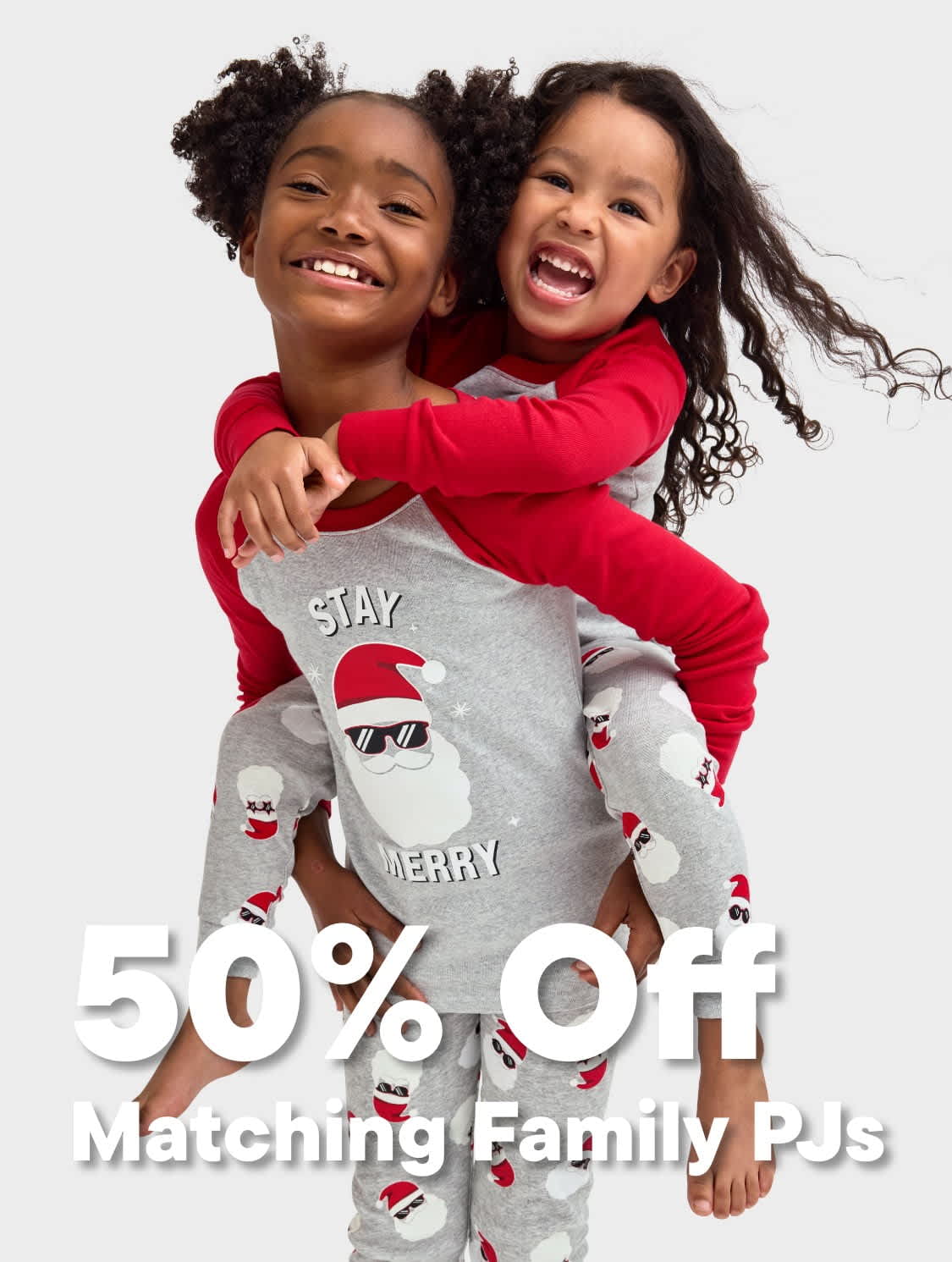 Two young girls with curly hair wearing matching red and gray pajamas are embracing and smiling joyfully against a plain white background.