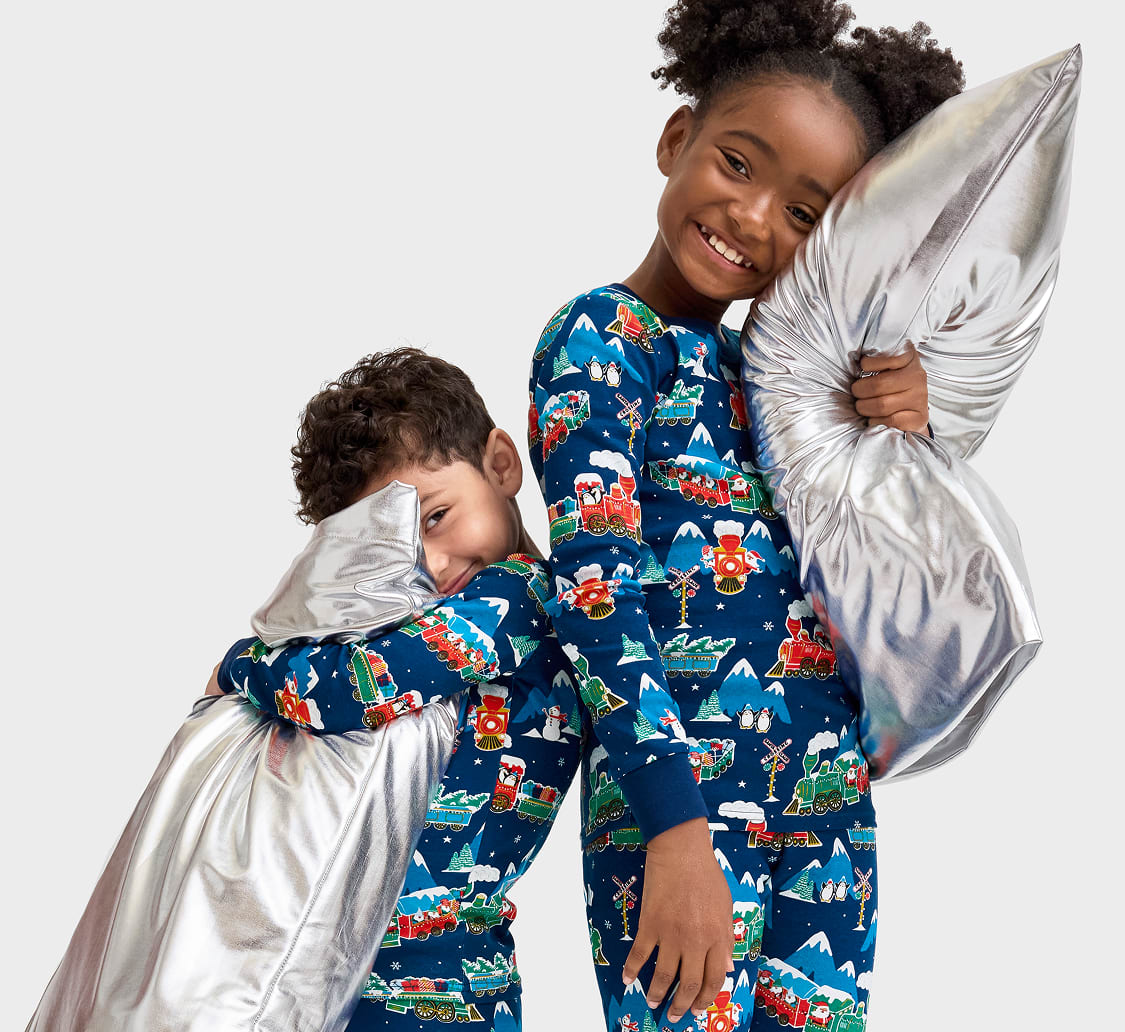 Two young children, a boy and a girl, are wearing matching pajamas with a colorful cartoon pattern and are smiling happily while holding a large, fluffy pillow.