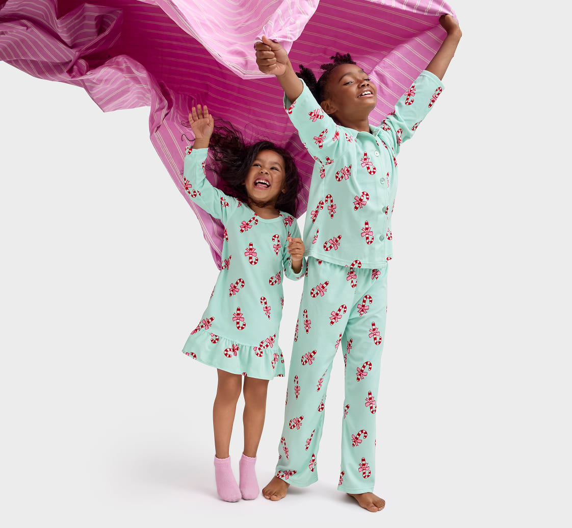 Two young girls in colorful, patterned pajamas are joyfully jumping and waving their arms, with a vibrant pink fabric billowing behind them.