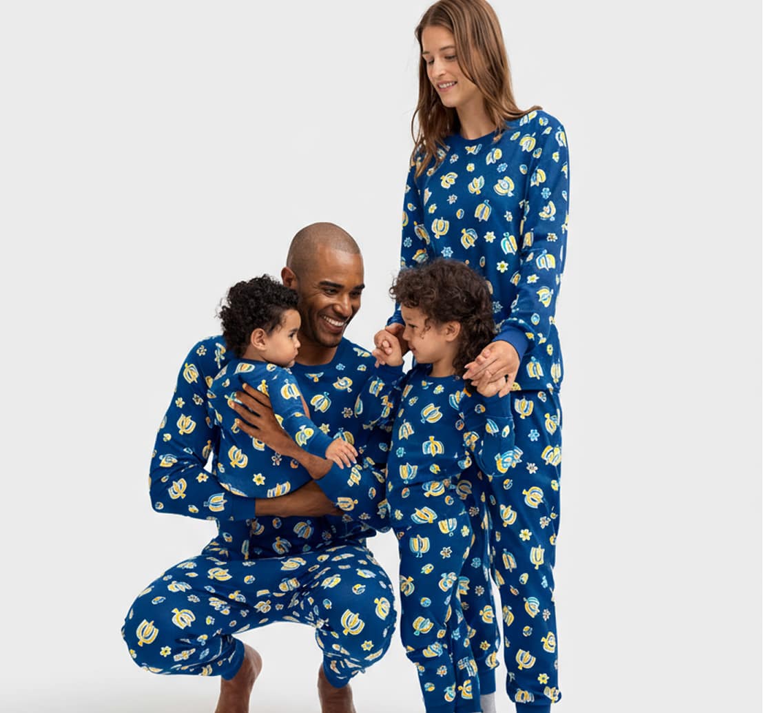 A family of four wearing matching blue pajamas with white daisy patterns, standing together and smiling against a plain white background with colorful shapes in the background.