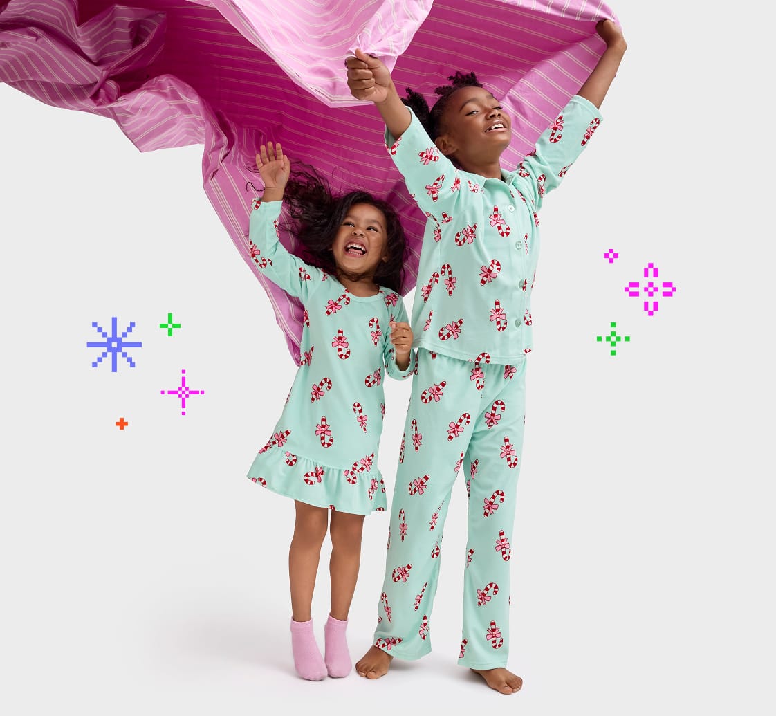 Two young girls in colorful, patterned pajamas are joyfully jumping and waving their arms, with a vibrant pink fabric billowing behind them.