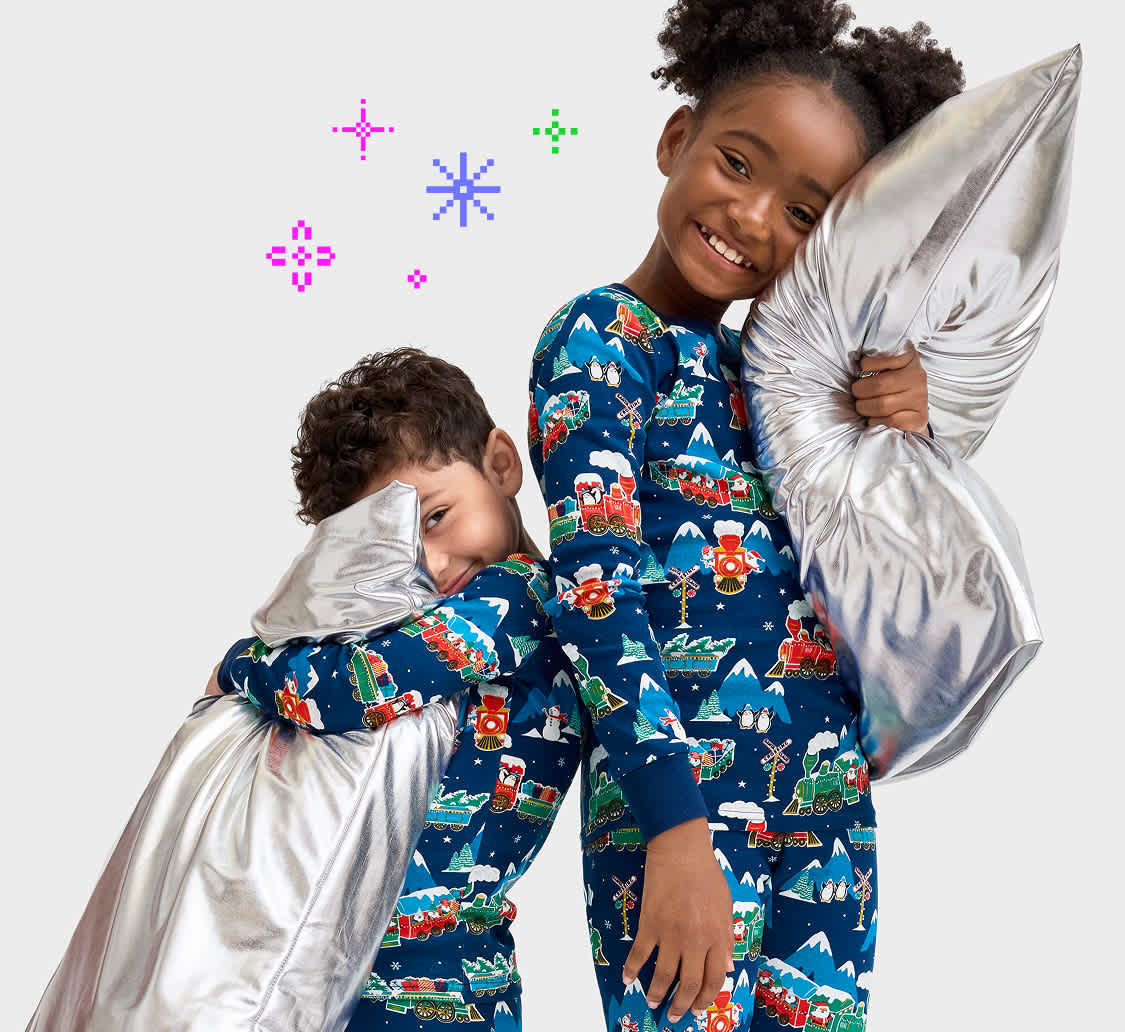 Two young children, a boy and a girl, are wearing matching pajamas with a colorful cartoon pattern and are smiling happily while holding a large, fluffy pillow.