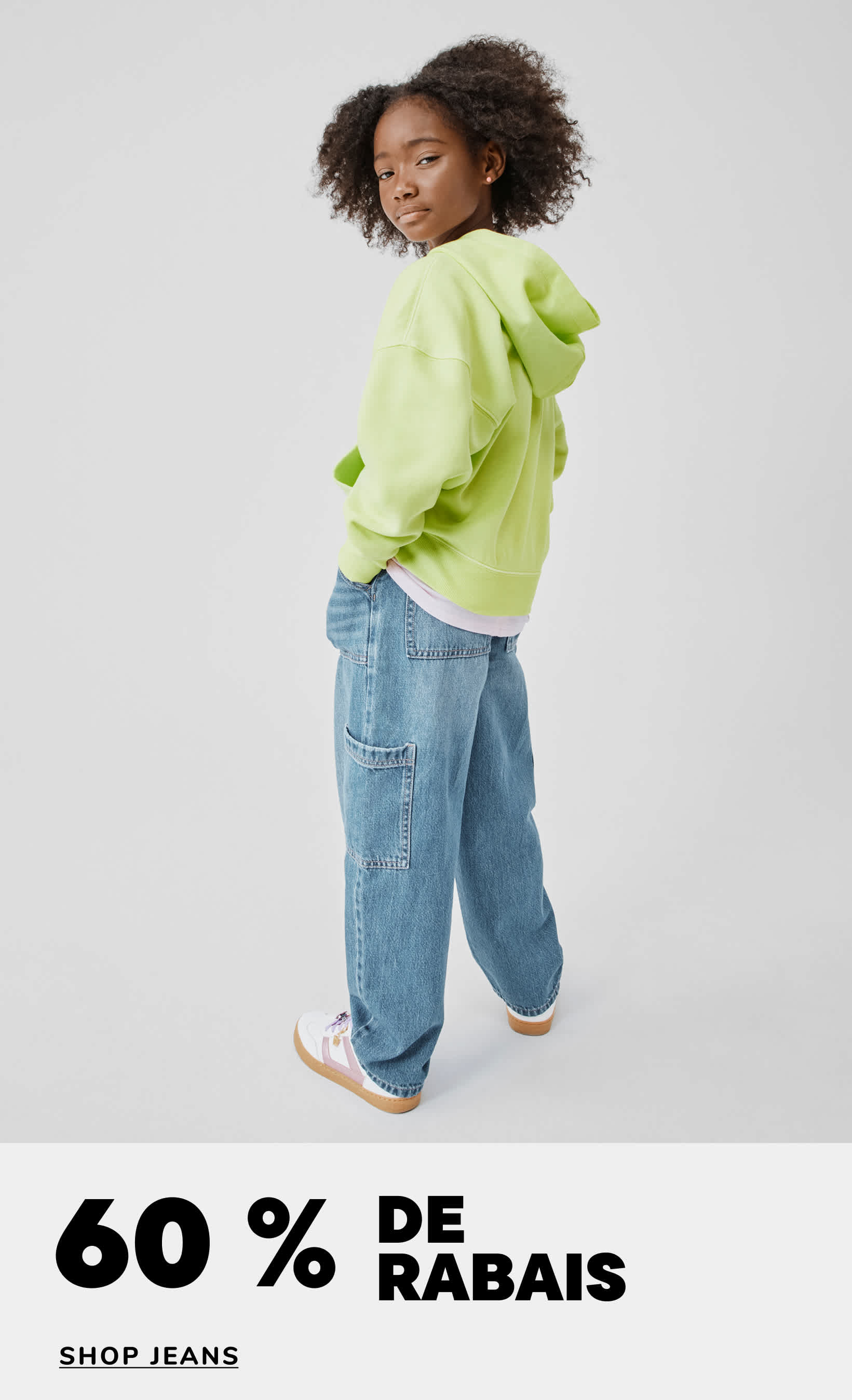 Une jeune personne aux cheveux bouclés, vêtue d'une doudoune vert vif et d'un jean ample, se détache sur un fond blanc uni, avec le texte « 60 % de réduction sur les jeans » en surimpression.