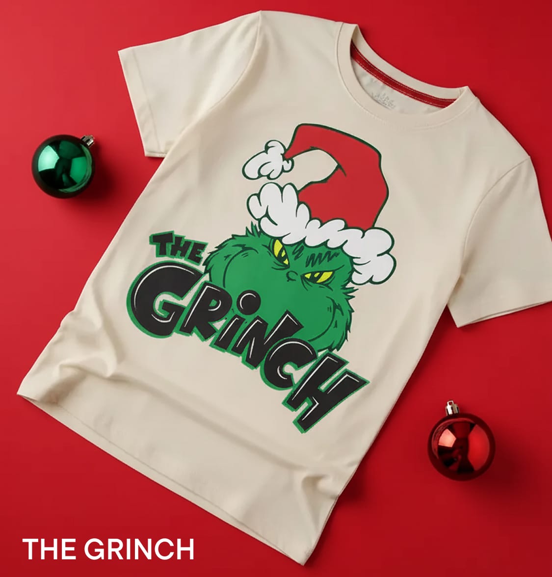 Un t-shirt blanc orné d'un motif graphique représentant le Grinch, personnage du célèbre conte de Noël, sur un fond rouge vif décoré de boules de Noël.