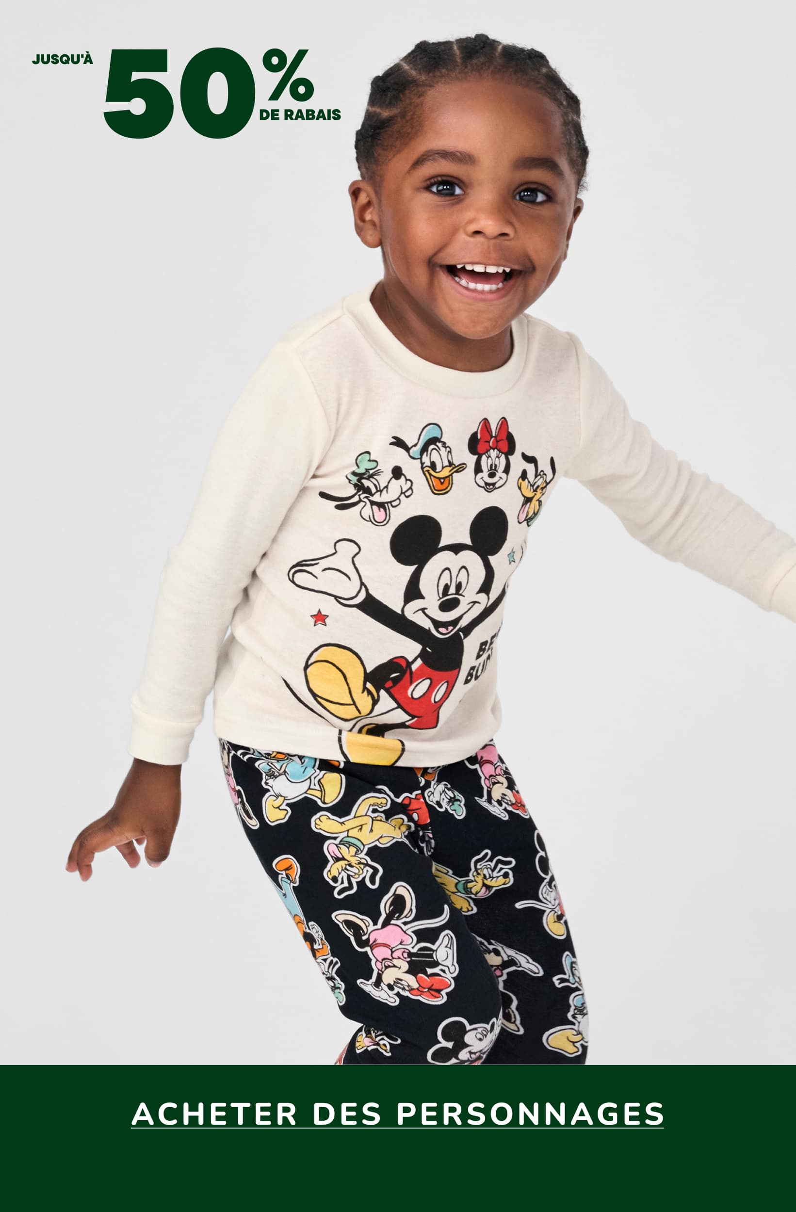 Un jeune enfant portant une chemise blanche à manches longues ornée d'un motif Mickey Mouse et un pantalon noir à motifs de dessins animés colorés, se tient devant un fond blanc uni avec une bannière verte annonçant une vente de produits dérivés à l'effigie de personnages.